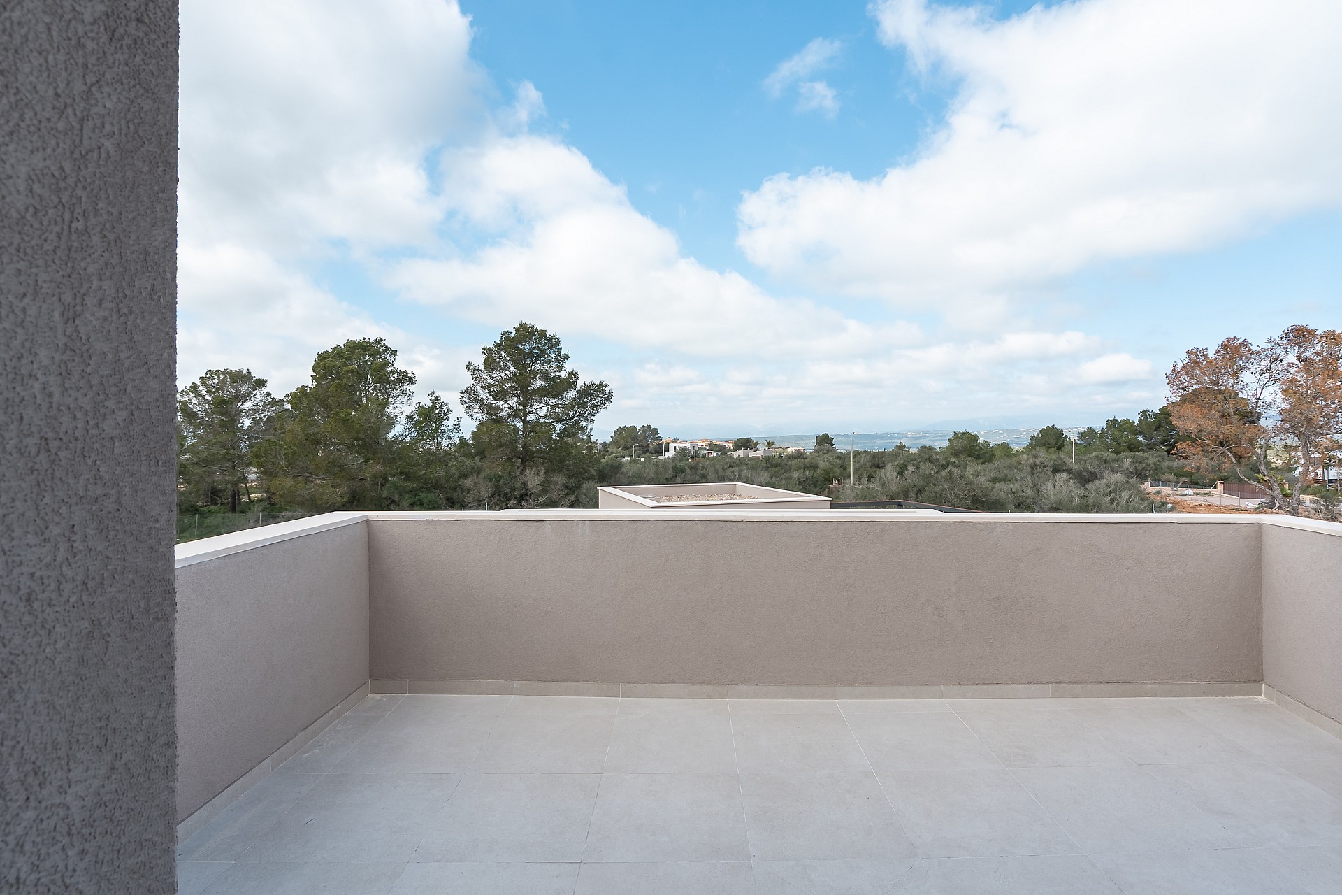 A wide, empty stone-tiled terrace with low perimeter walls, overlooking green trees and distant rolling hills under a blue sky.