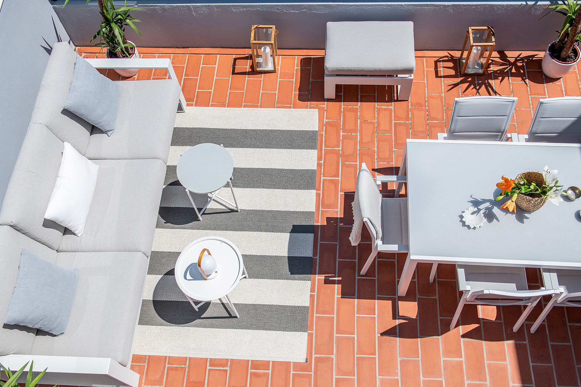 Aerial view of a tiled penthouse terrace featuring a gray sectional sofa, a white dining table with chairs, and a striped rug.