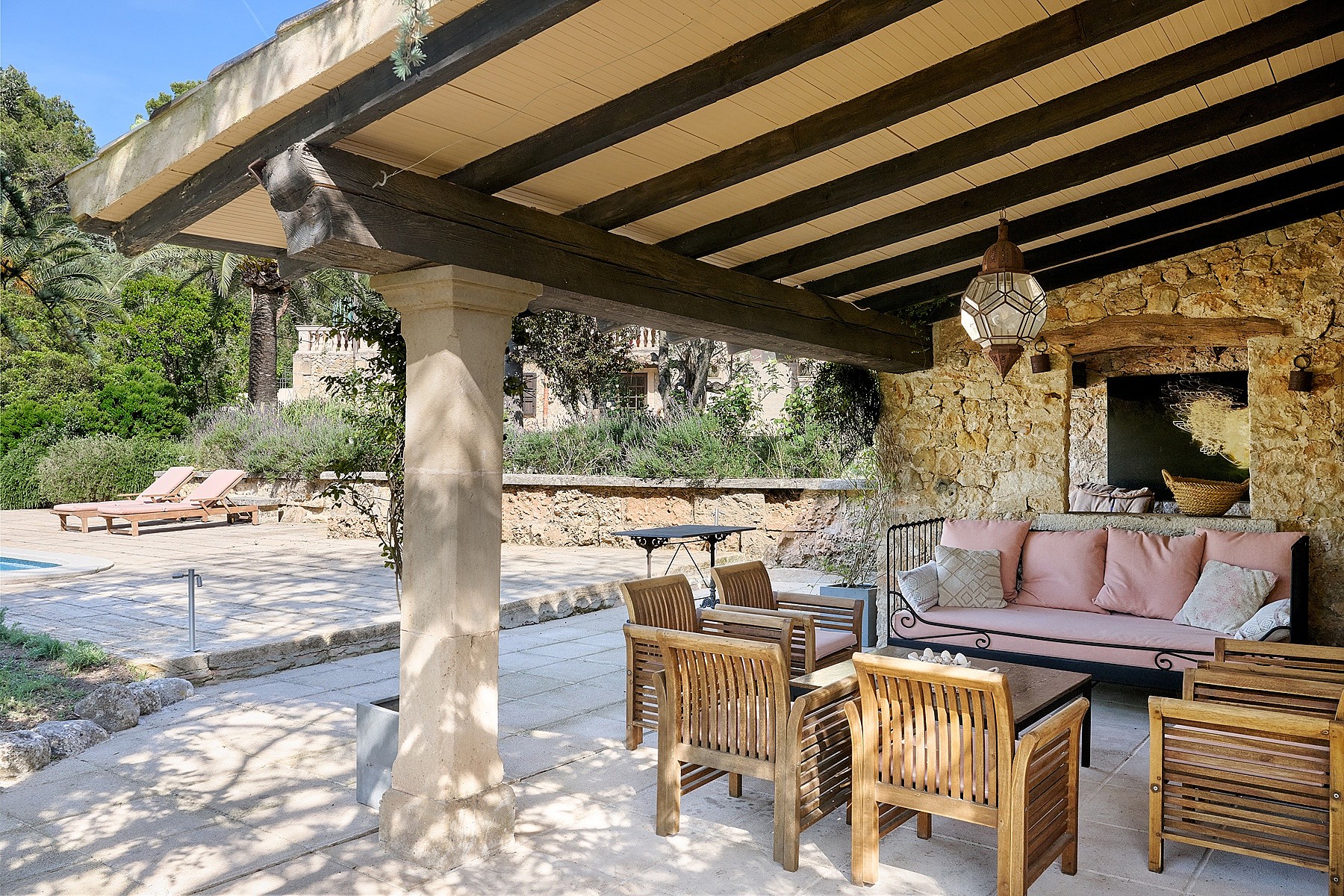 A shaded outdoor patio with exposed timber beams, stone walls, and wooden furniture overlooking a sunlit garden area.