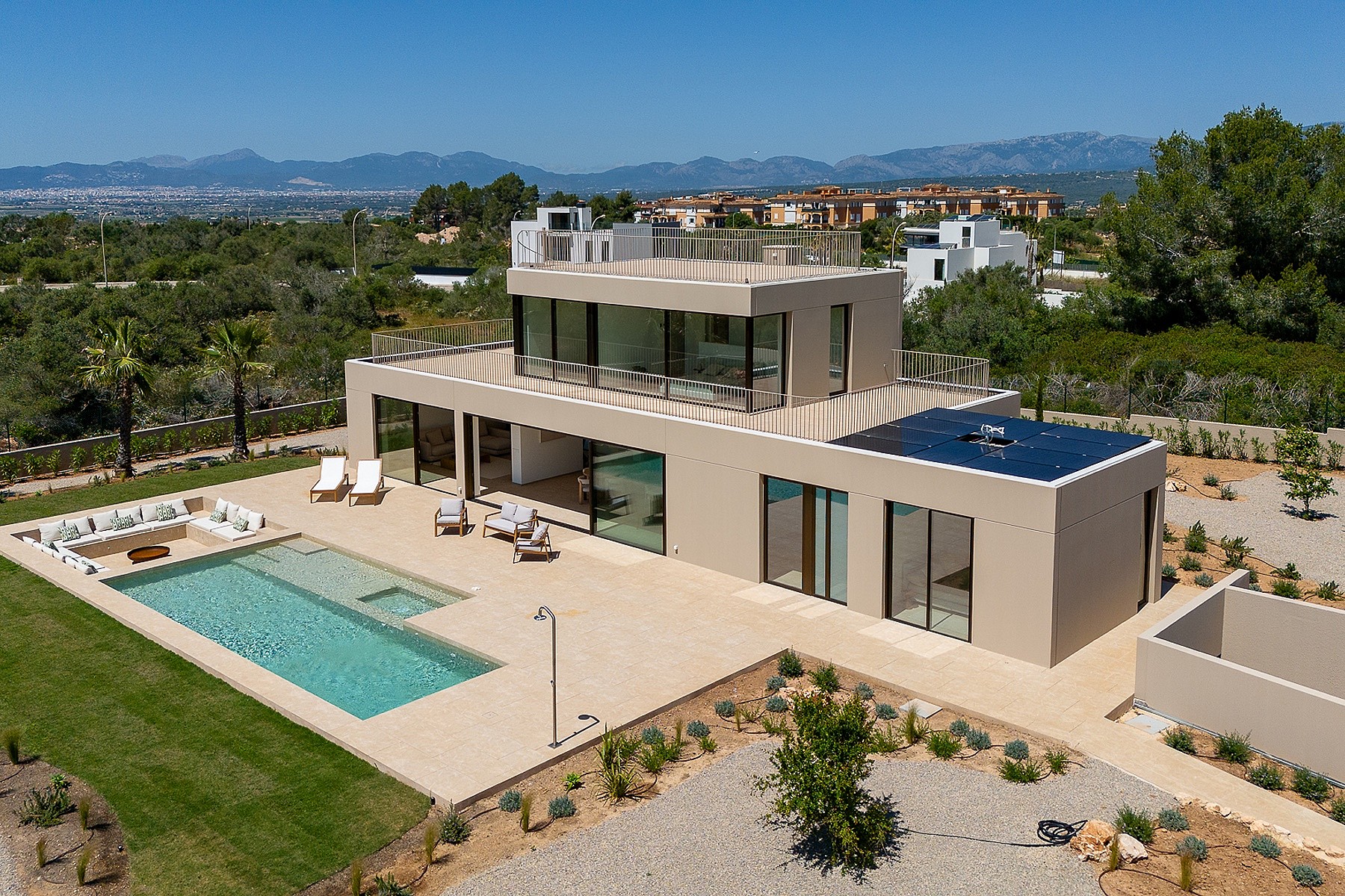 Aerial shot of a beige modern villa with a rectangular swimming pool, terrace seating, and a flat rooftop, set against mountain vistas.