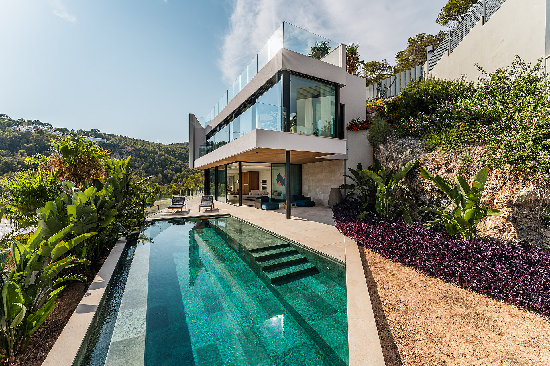 Modern two-story villa with glass walls and stone terracing, set against a rocky hill with an infinity-edge swimming pool.