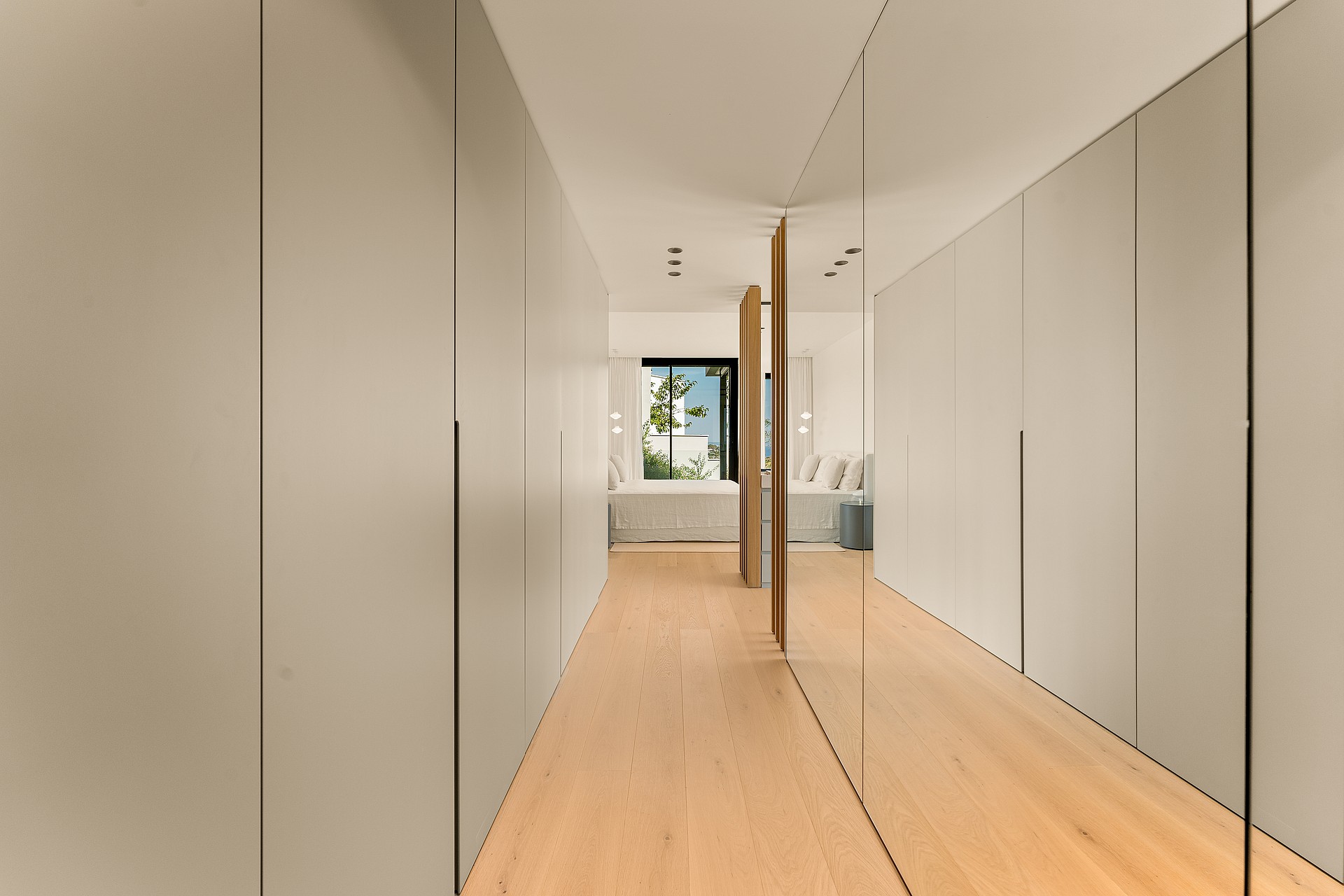 A corridor-style dressing room featuring full-height matte beige wardrobes and a large mirrored wall leading to a bright bedroom.