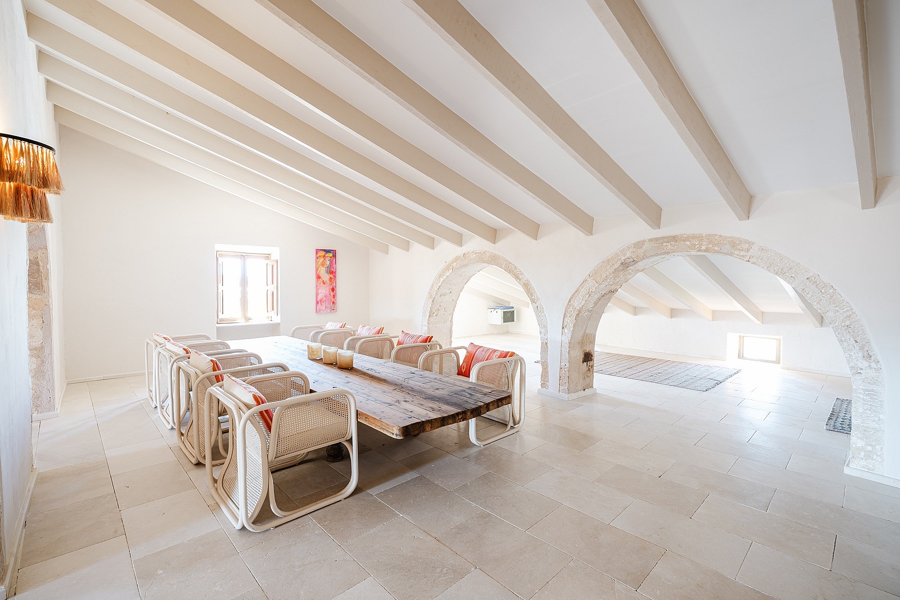 Spacious room with exposed wooden ceiling beams, stone floor, and arched partitions. Long rustic table with woven chairs.
