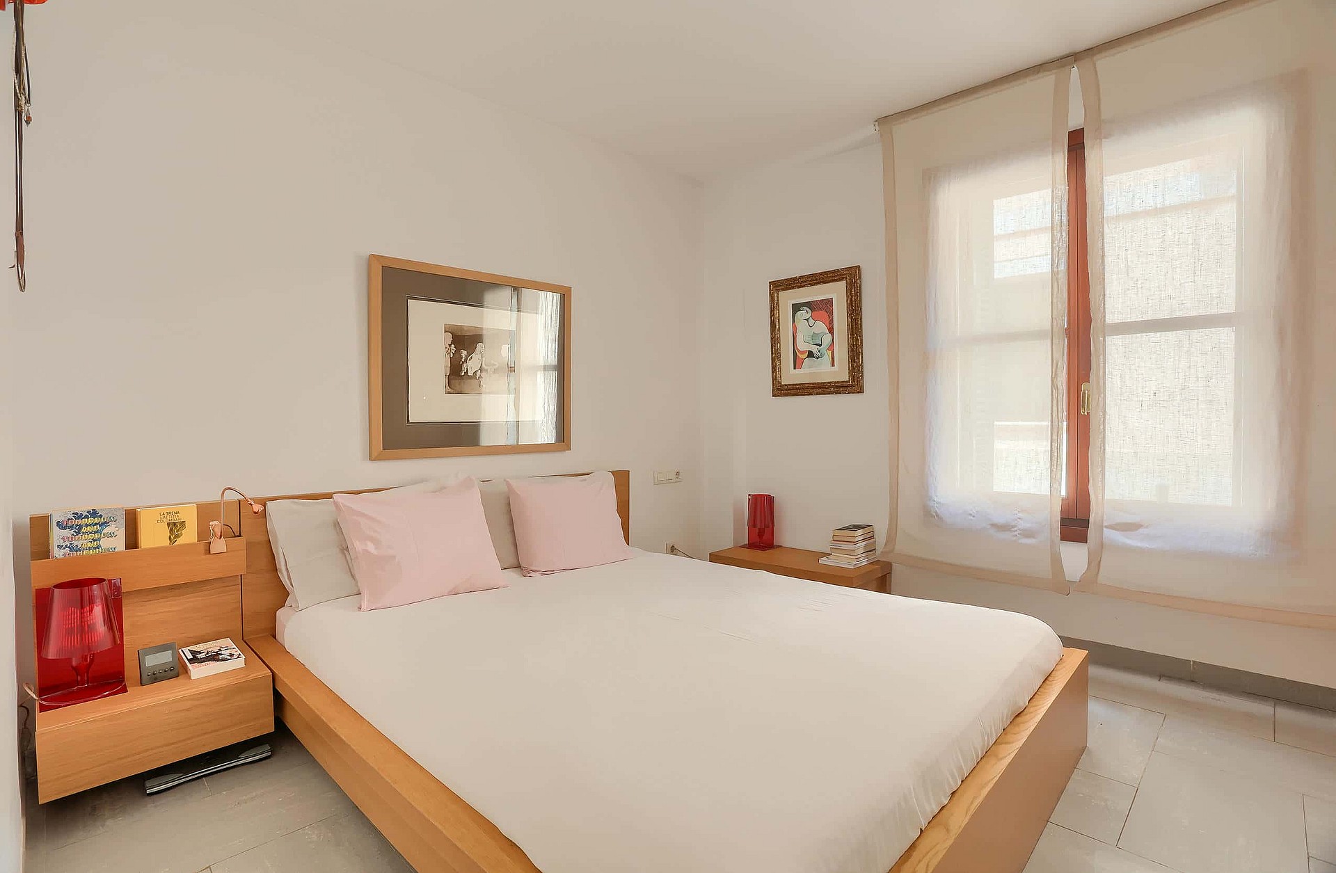 A double bedroom with light wooden bed frame, bedside storage units, and a window with sheer curtains.