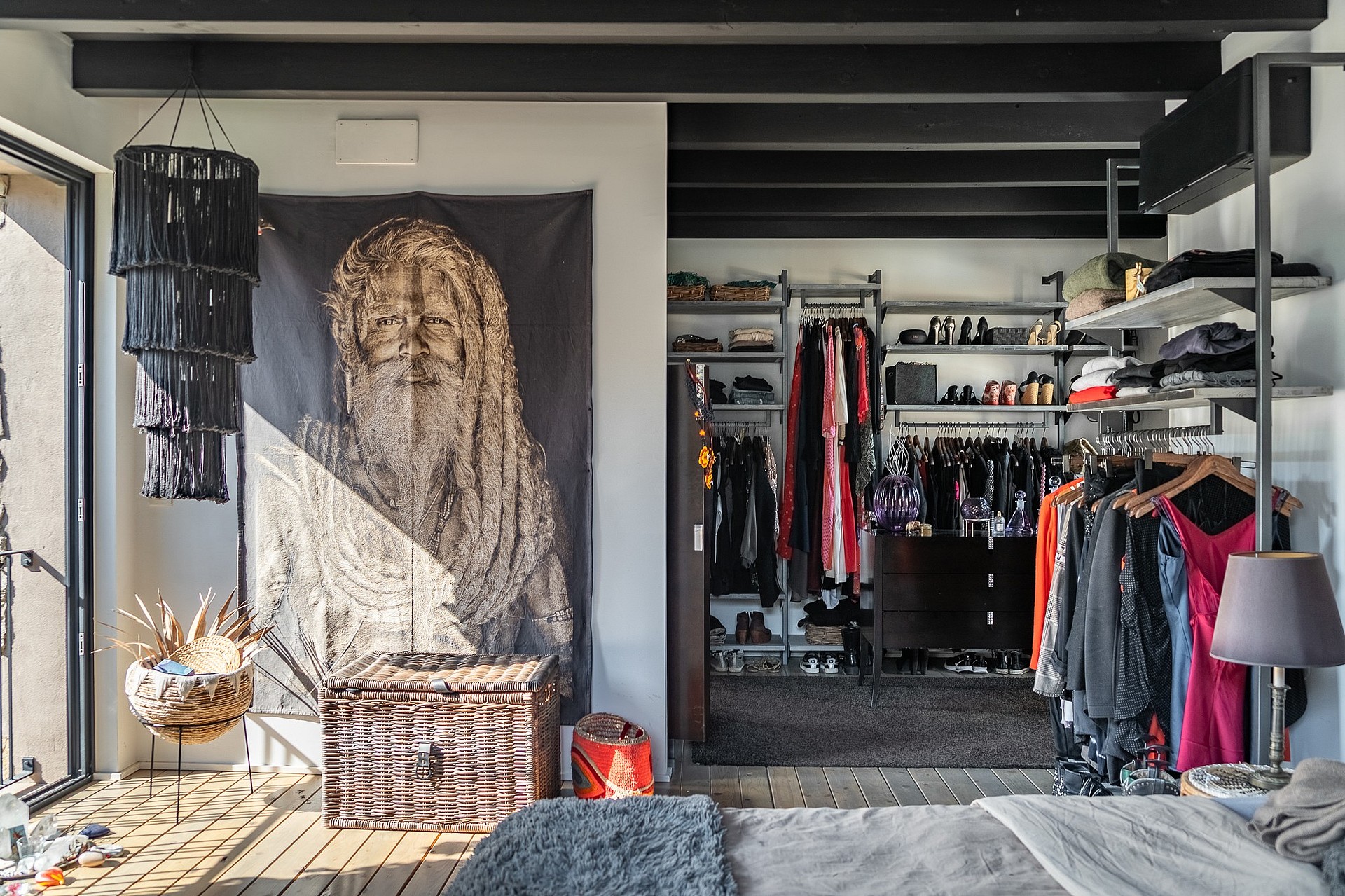 Walk-in wardrobe with industrial open shelving, dark wooden trunk, and decorative wall tapestry, illuminated by natural light.