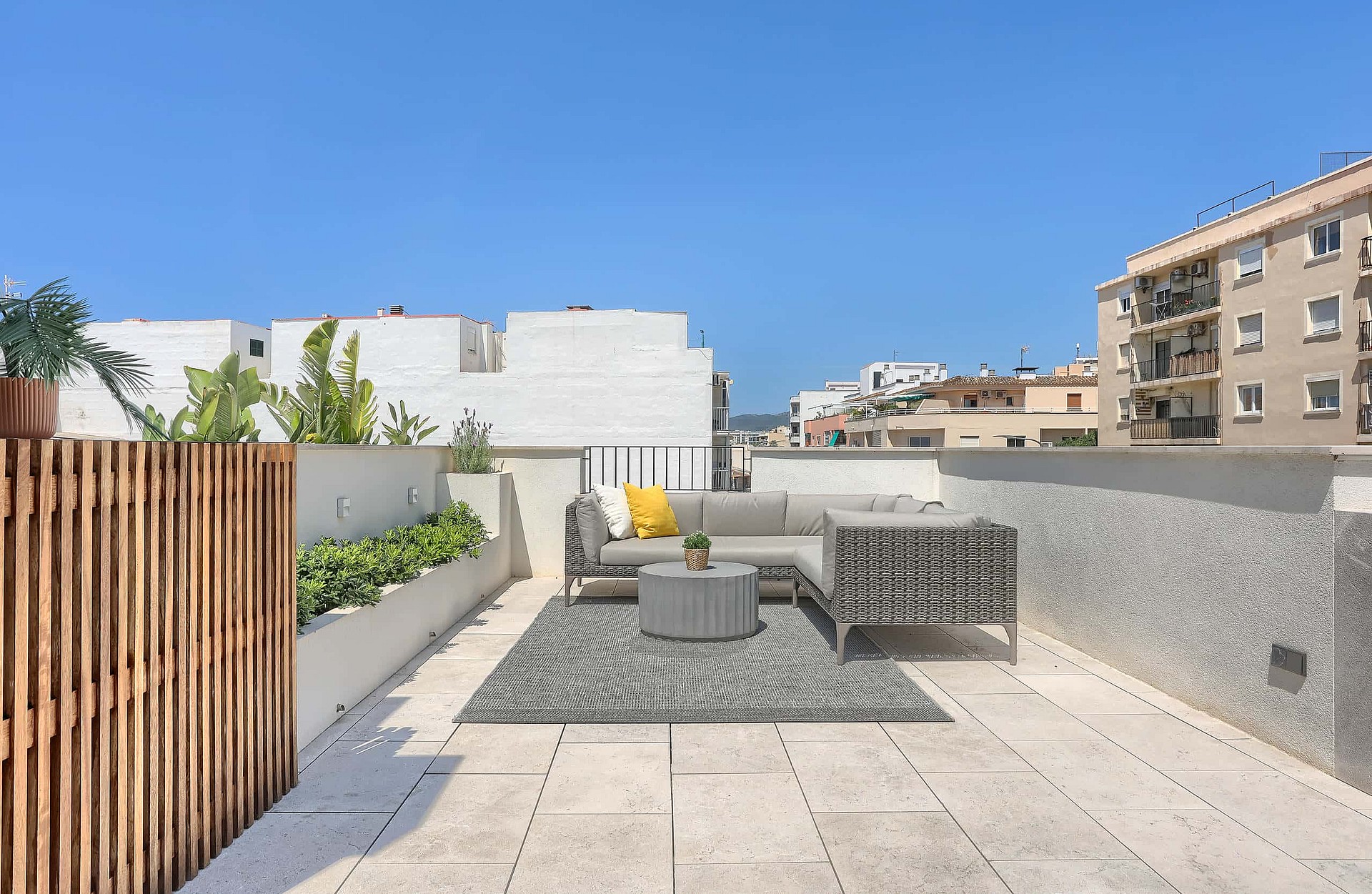 A sunny rooftop terrace featuring light stone flooring, a modular grey sofa, patterned rug, and a wood-slatted privacy screen.