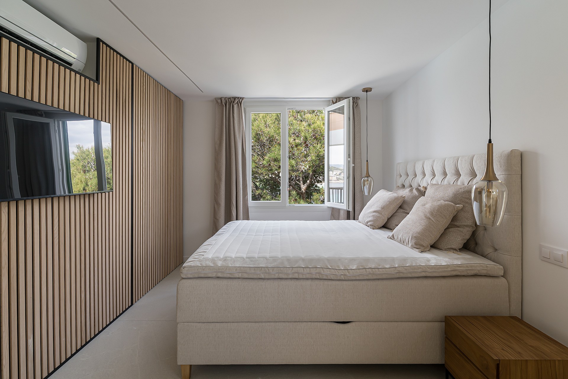 Master bedroom featuring vertical light wood wall panels, a wall-mounted television, and windows overlooking green trees.