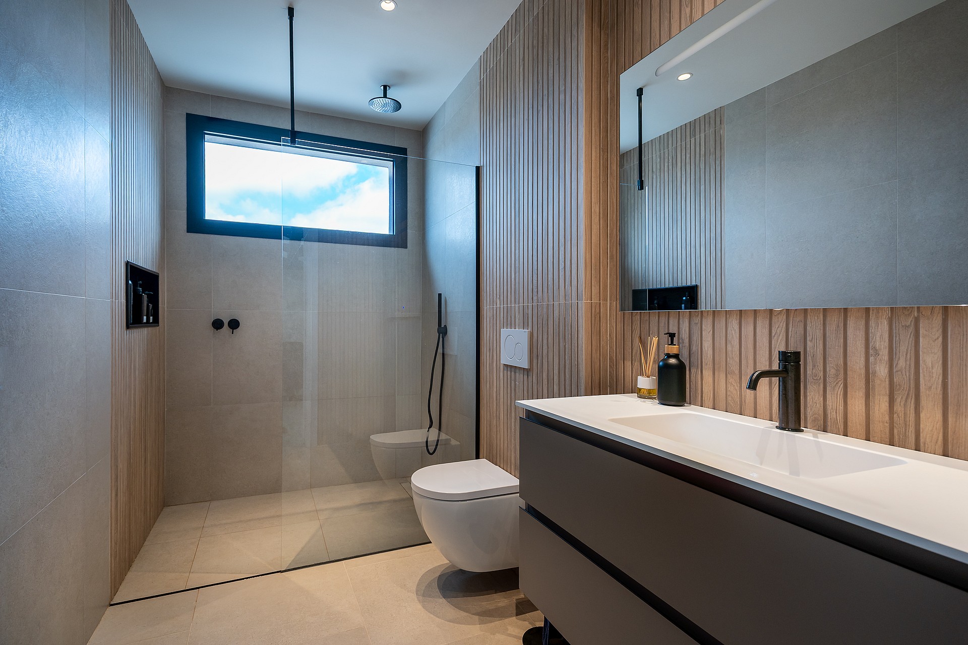 A modern bathroom featuring a floating grey vanity, wood slat wall panels, walk-in shower, and a minimalist white toilet.