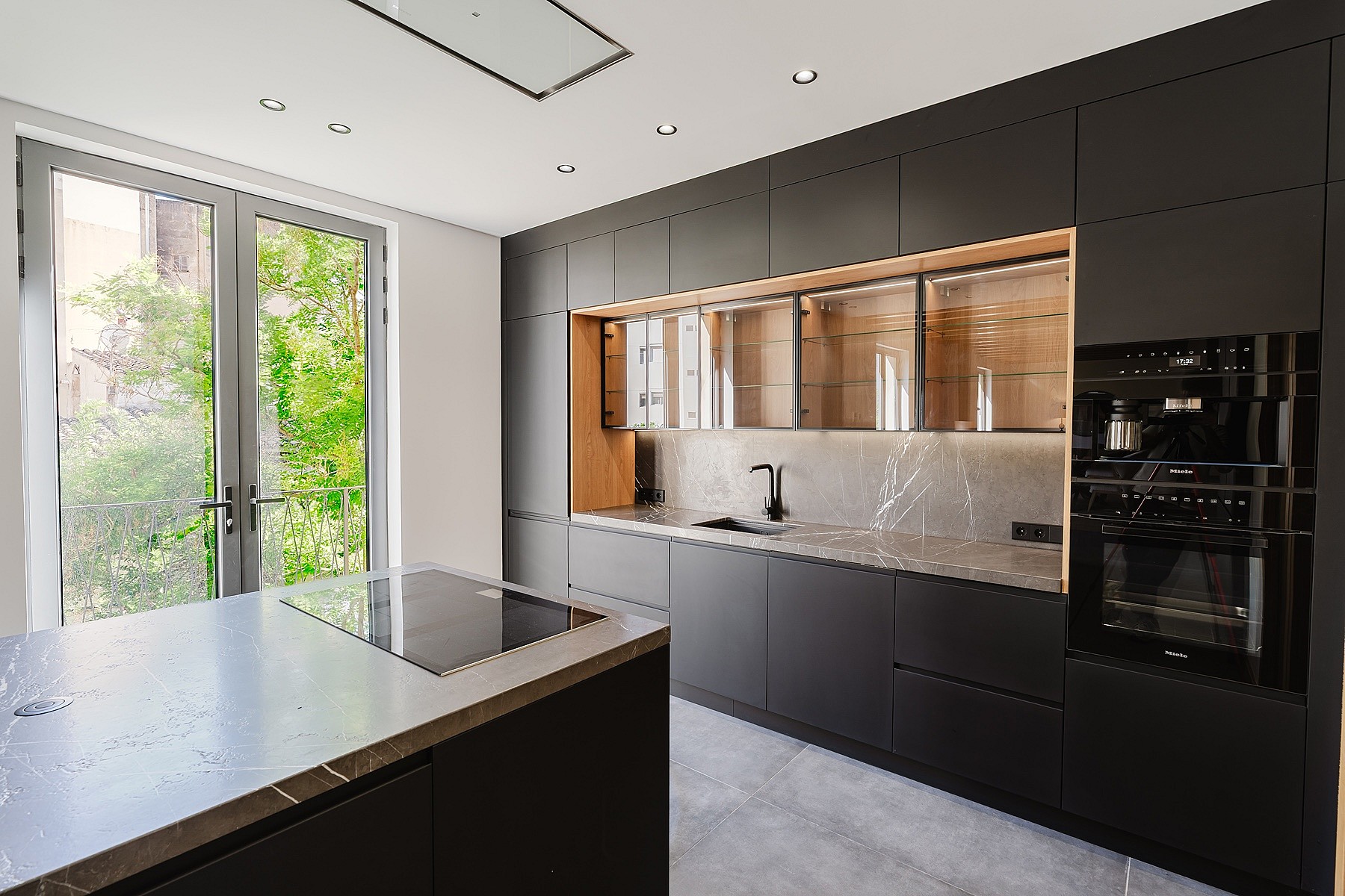 A modern kitchen with matte black cabinetry, a central island with an integrated hob, and French doors opening to greenery.