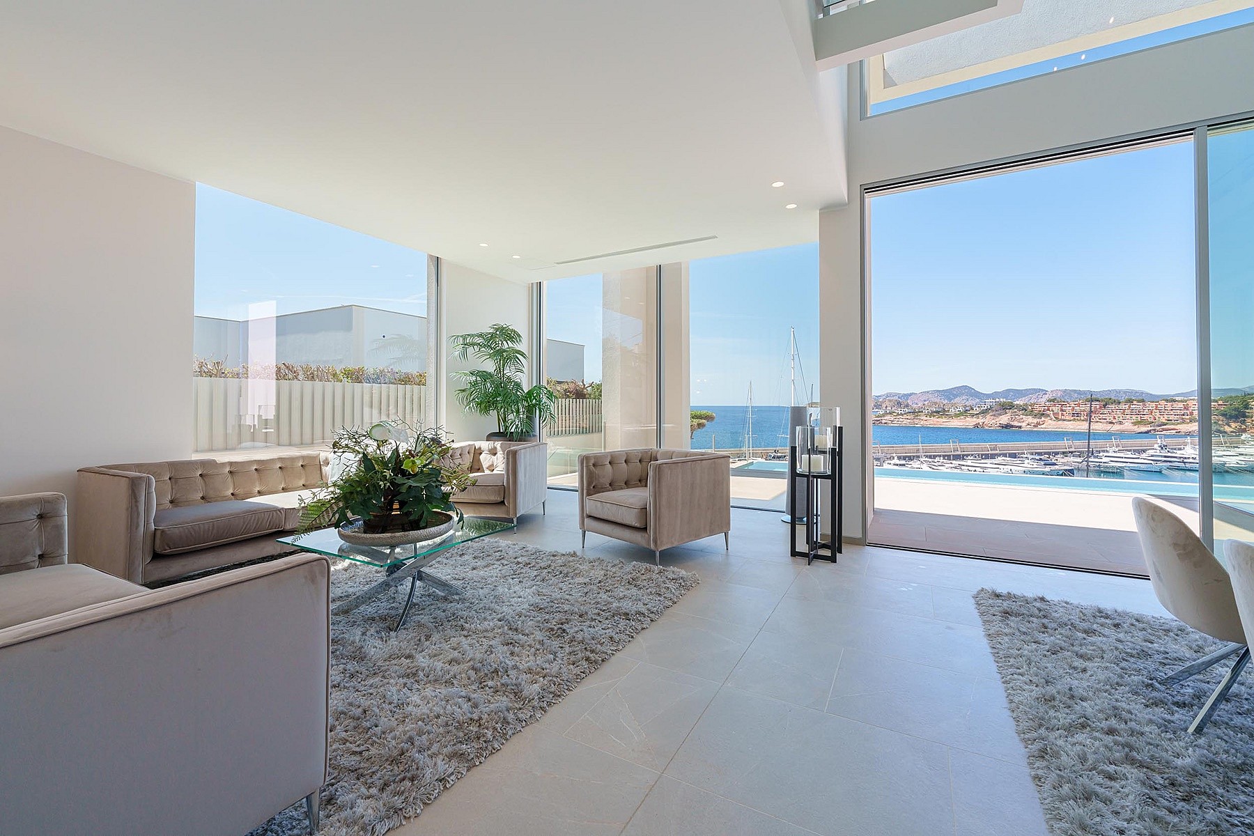 Bright living room with white walls, tufted beige sofas, glass coffee table, and large windows looking out to the harbor.
