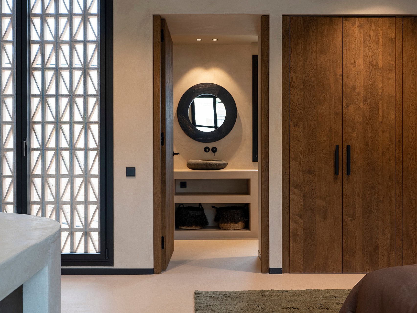 View from a bedroom into a modern bathroom with a circular mirror, stone basin, and wooden wardrobe doors on the right.