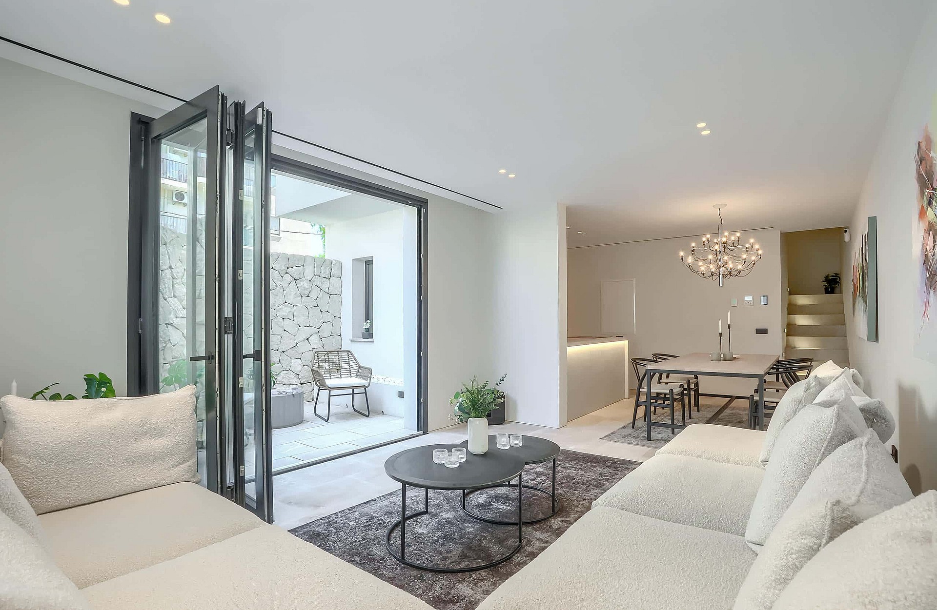 A modern living room with neutral fabric sofas, a coffee table, and folding glass doors leading to a stone-walled terrace.