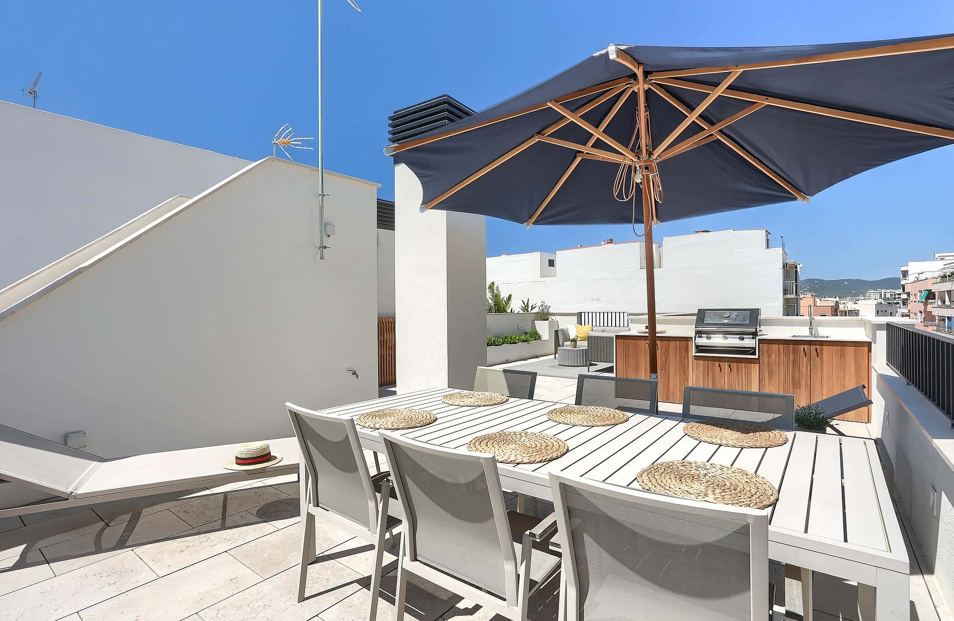 A sunny rooftop terrace in El Molinar featuring a white dining table with woven mats, a large navy umbrella, and built-in grill.