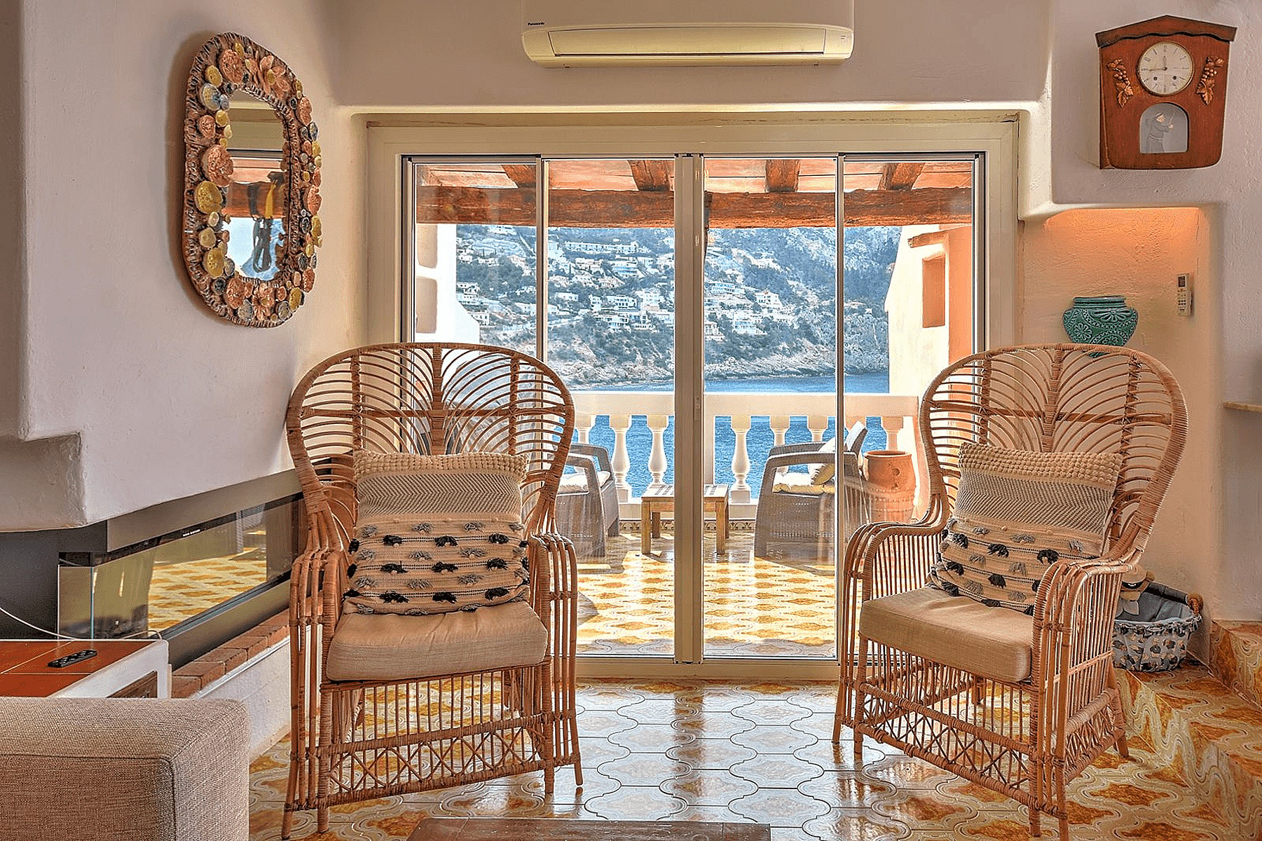 Two rattan armchairs facing sliding glass doors that open onto a sea-view terrace, with decorative mirrors and wall clock.