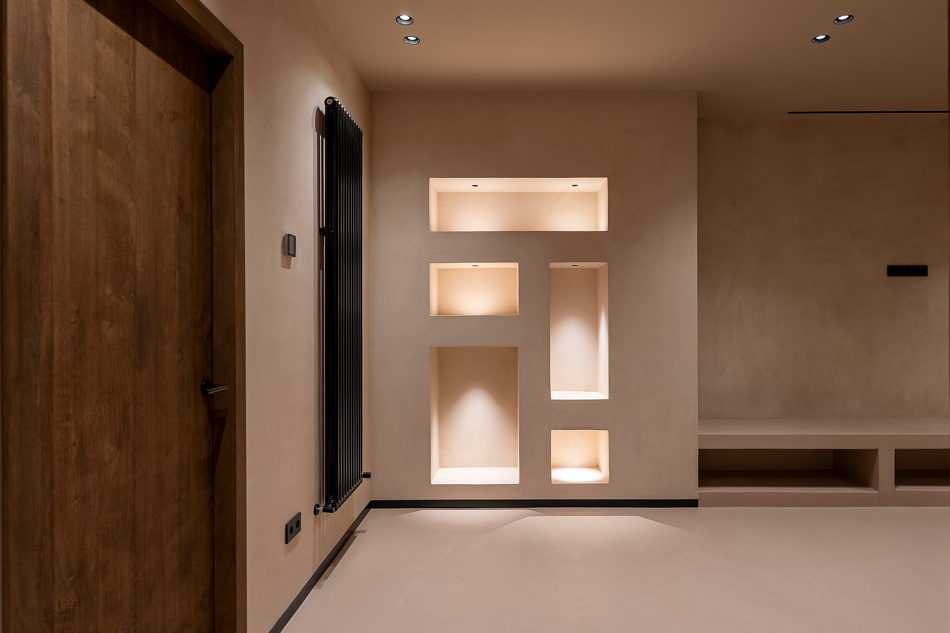 A neutral-toned living space featuring recessed wall shelving with internal warm lighting, a natural wood door, and a low bench.