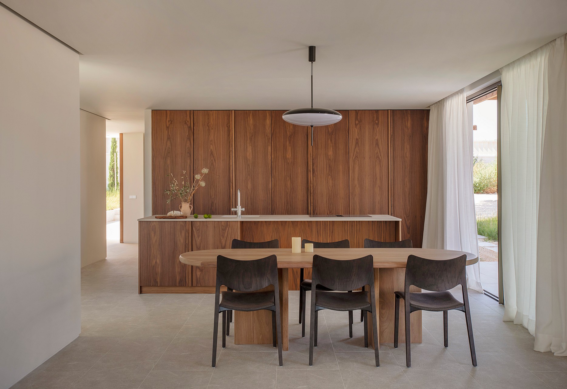 A minimalist dining area featuring an oval wood table and dark chairs, set against full-height walnut kitchen cabinetry.