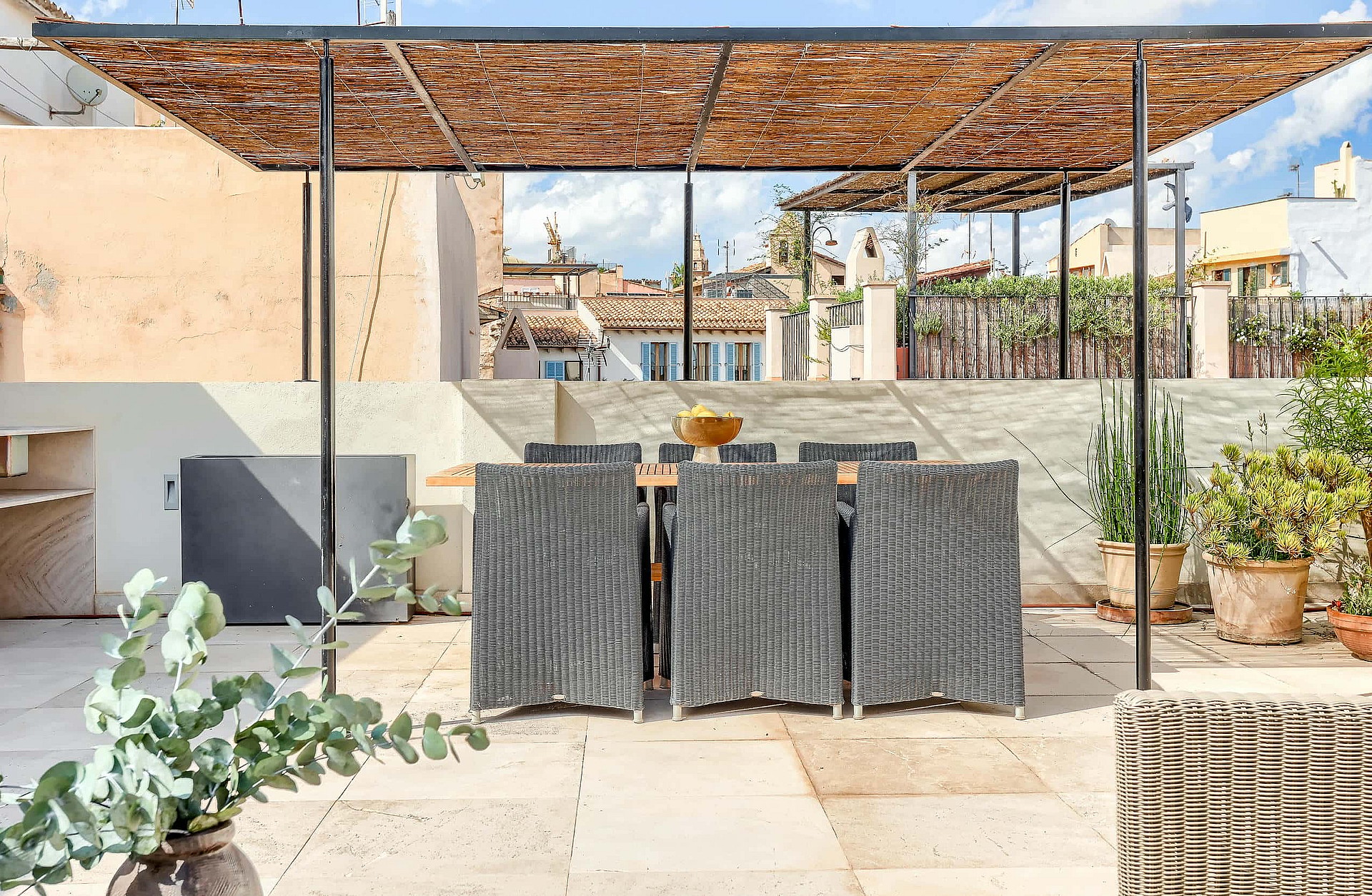 A rooftop terrace dining space with a dark metal pergola, woven chairs, and a view of old town buildings under a blue sky.