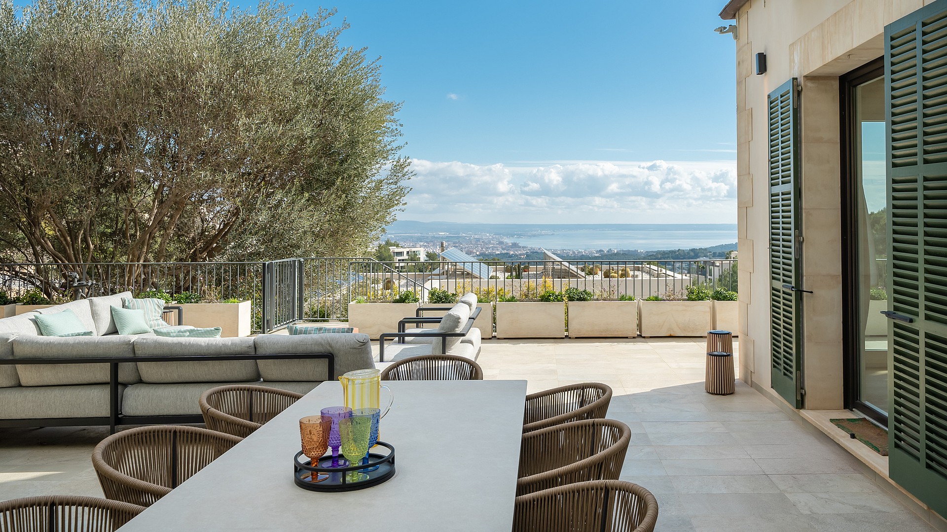Outdoor dining terrace featuring a stone table, woven chairs, and a large olive tree overlooking the Son Vida landscape.