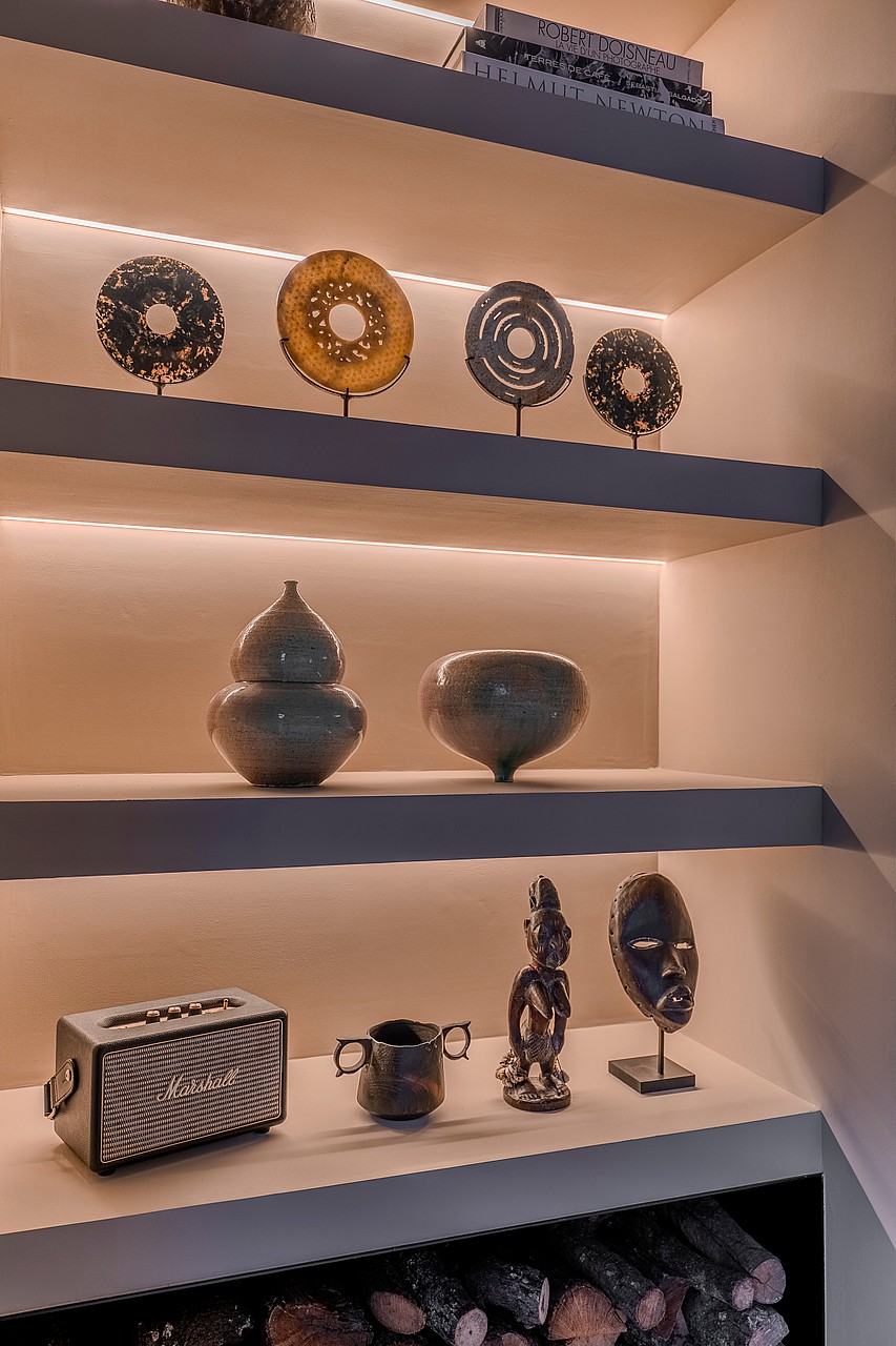 Floating shelves display ceramic art and decorative masks under integrated LED strips above a firewood storage area.