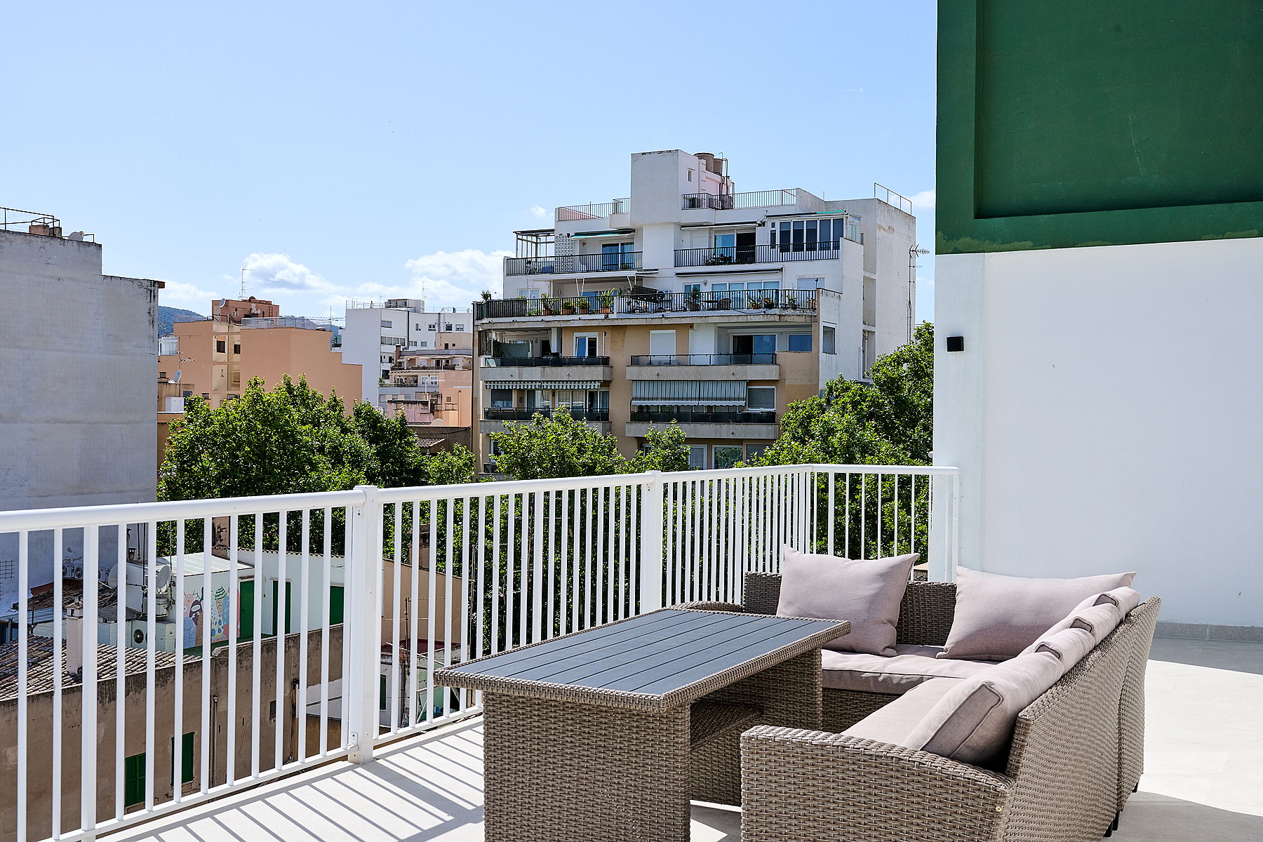 A sunny outdoor terrace with rattan-style sofa seating, white metal railings, and a view of urban residential buildings.