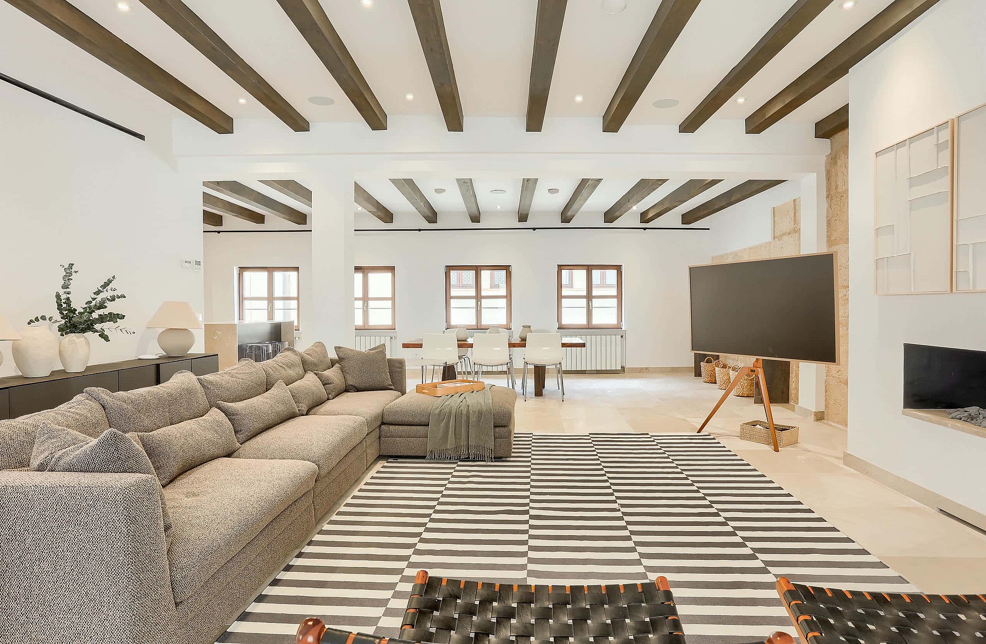 A spacious living room featuring exposed dark wood ceiling beams, white walls, and a large patterned sectional sofa on a striped rug.