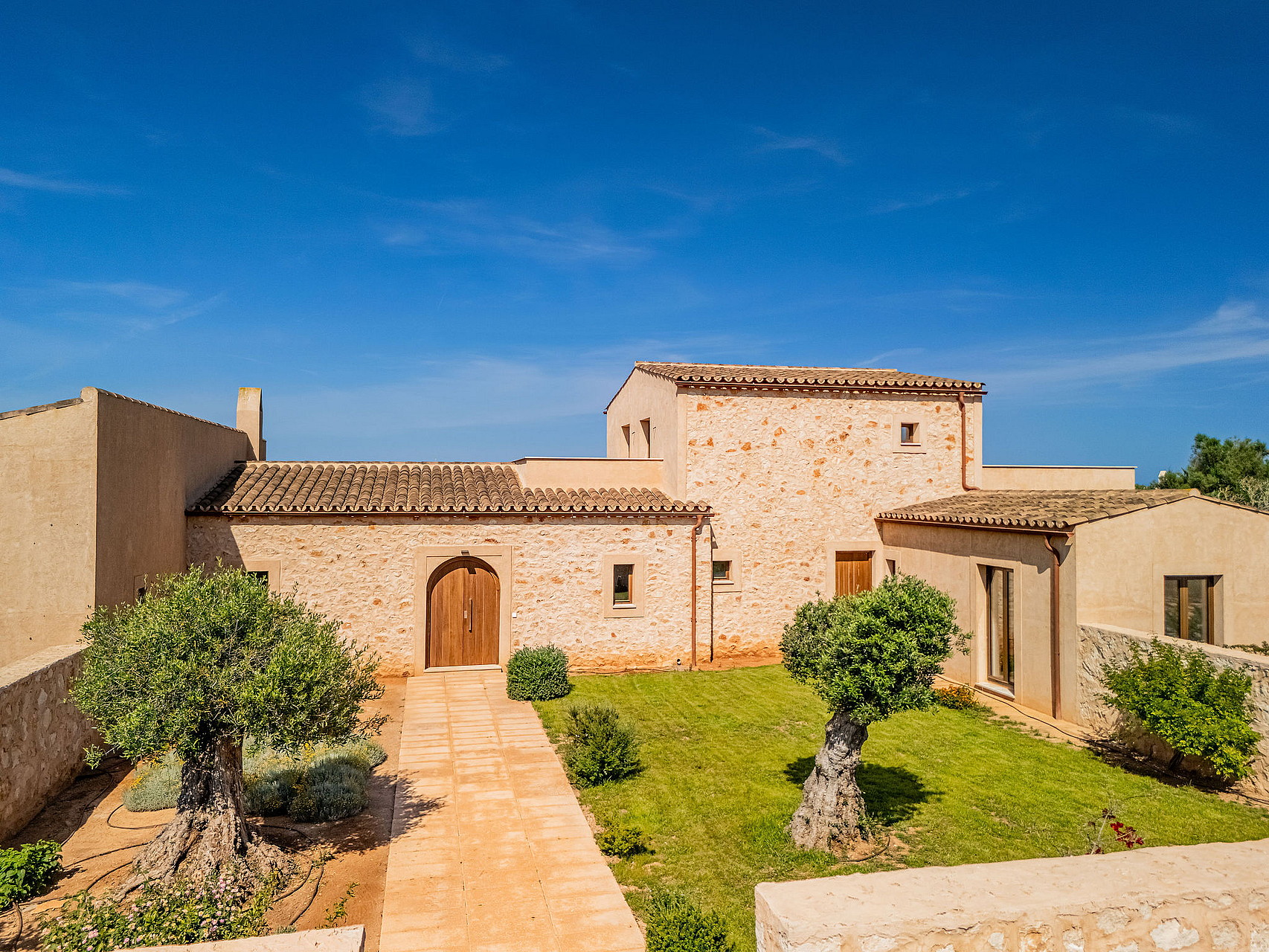 Exterior view of a traditional stone finca in Santanyí with a paved pathway leading to a wooden arched door, surrounded by a lawn and olive trees under a bright blue sky.