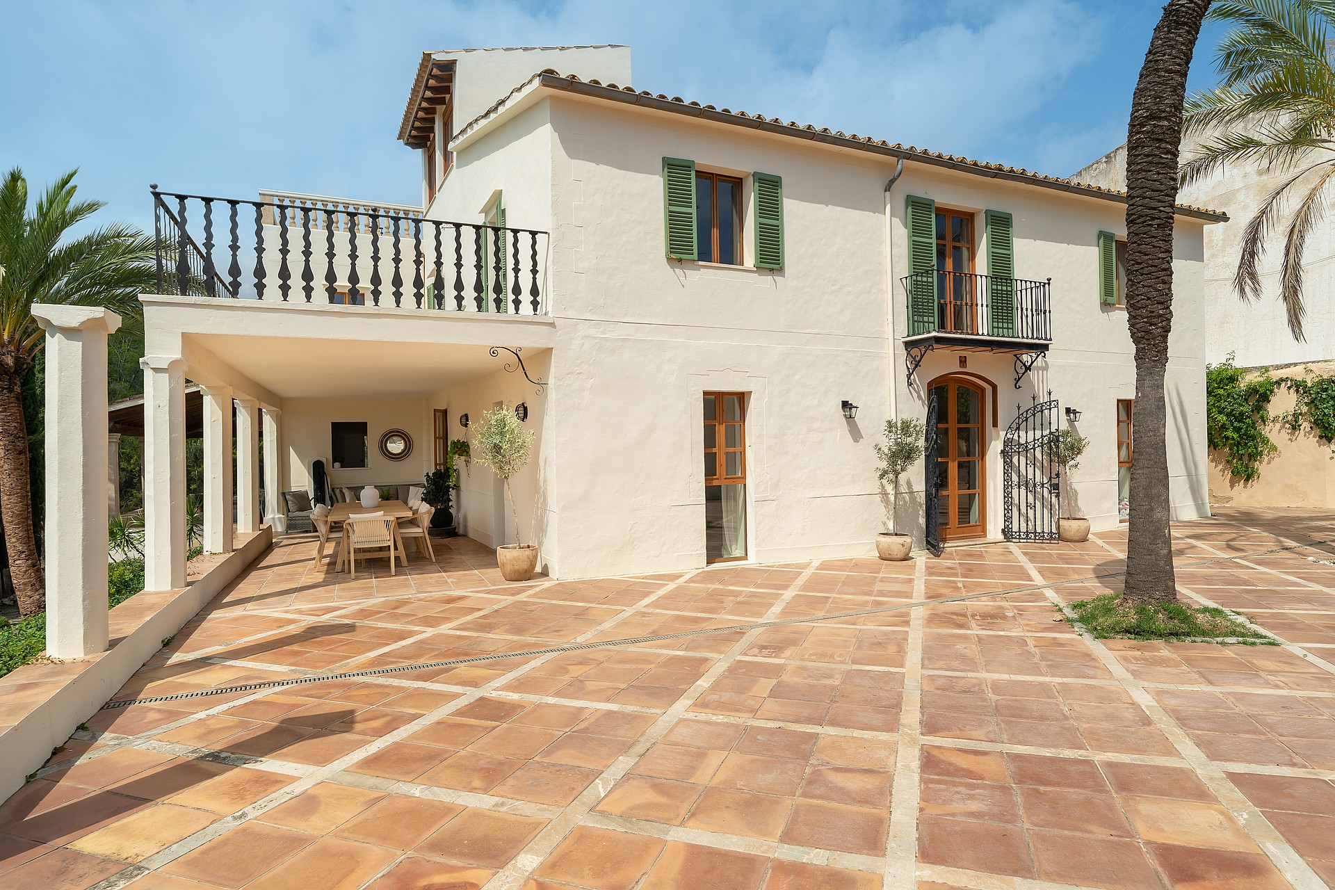 Two-story sand-colored villa with green shutters, a large terracotta-tiled patio, and a covered dining area.