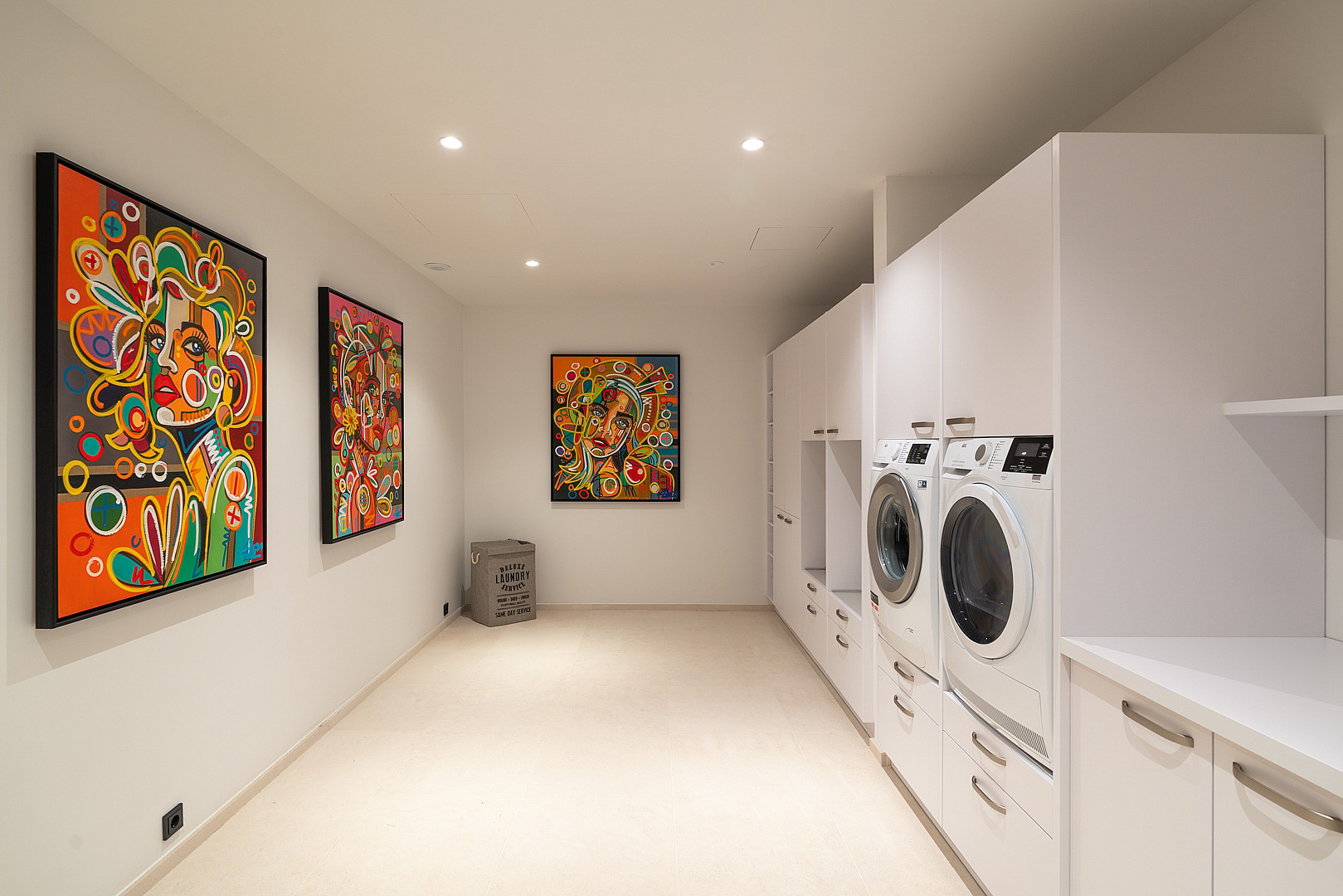 A bright, clean laundry room with white cabinetry, integrated appliances, and three vibrant abstract paintings on the wall.