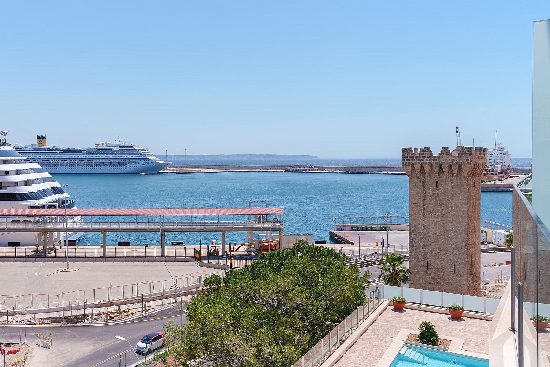 Vista elevada sobre un puerto con una torre histórica, cruceros y el mar bajo un cielo despejado.