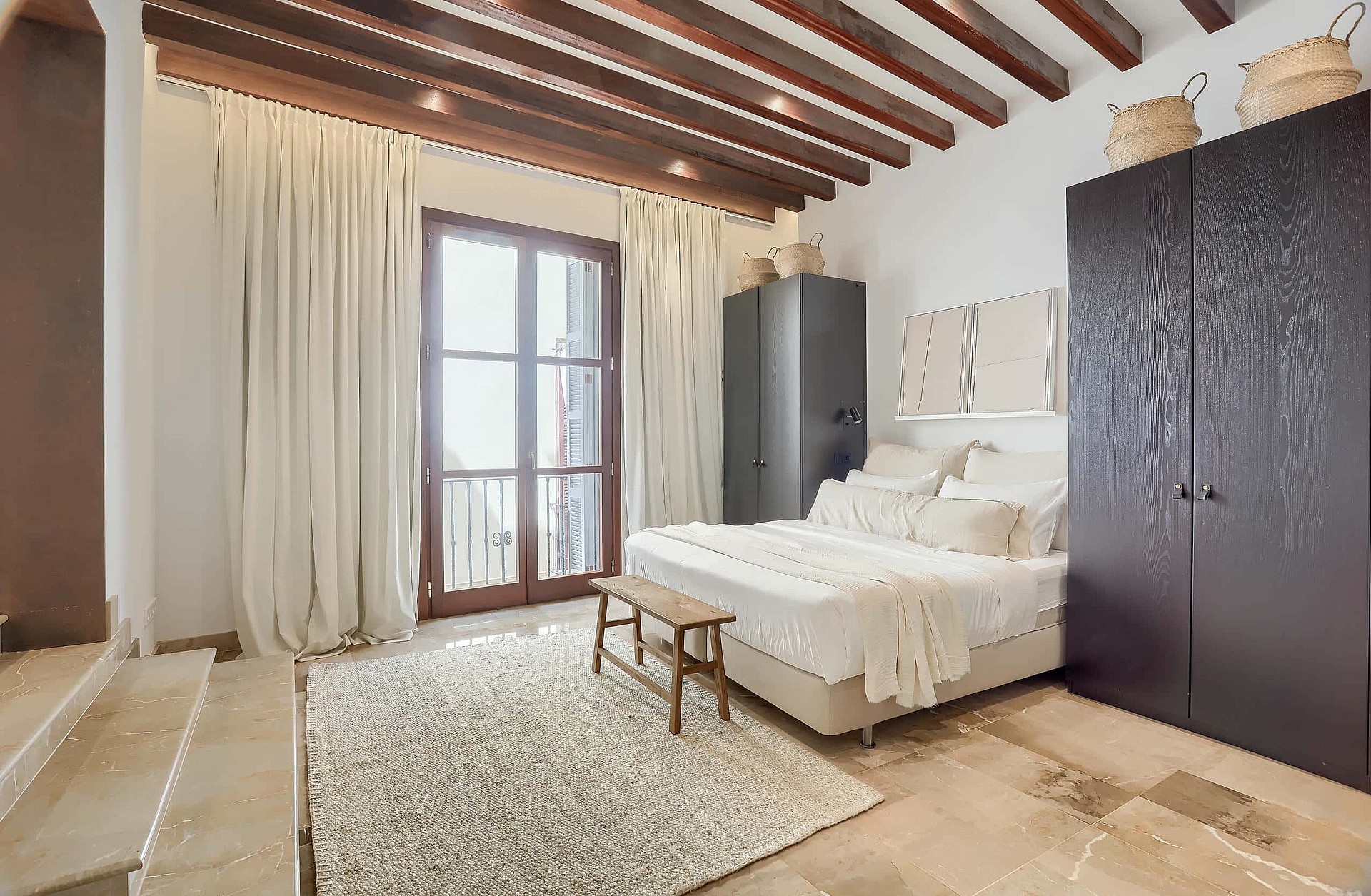 A bright bedroom featuring exposed dark wooden ceiling beams, cream drapes, a beige bed, and dark wood wardrobes.