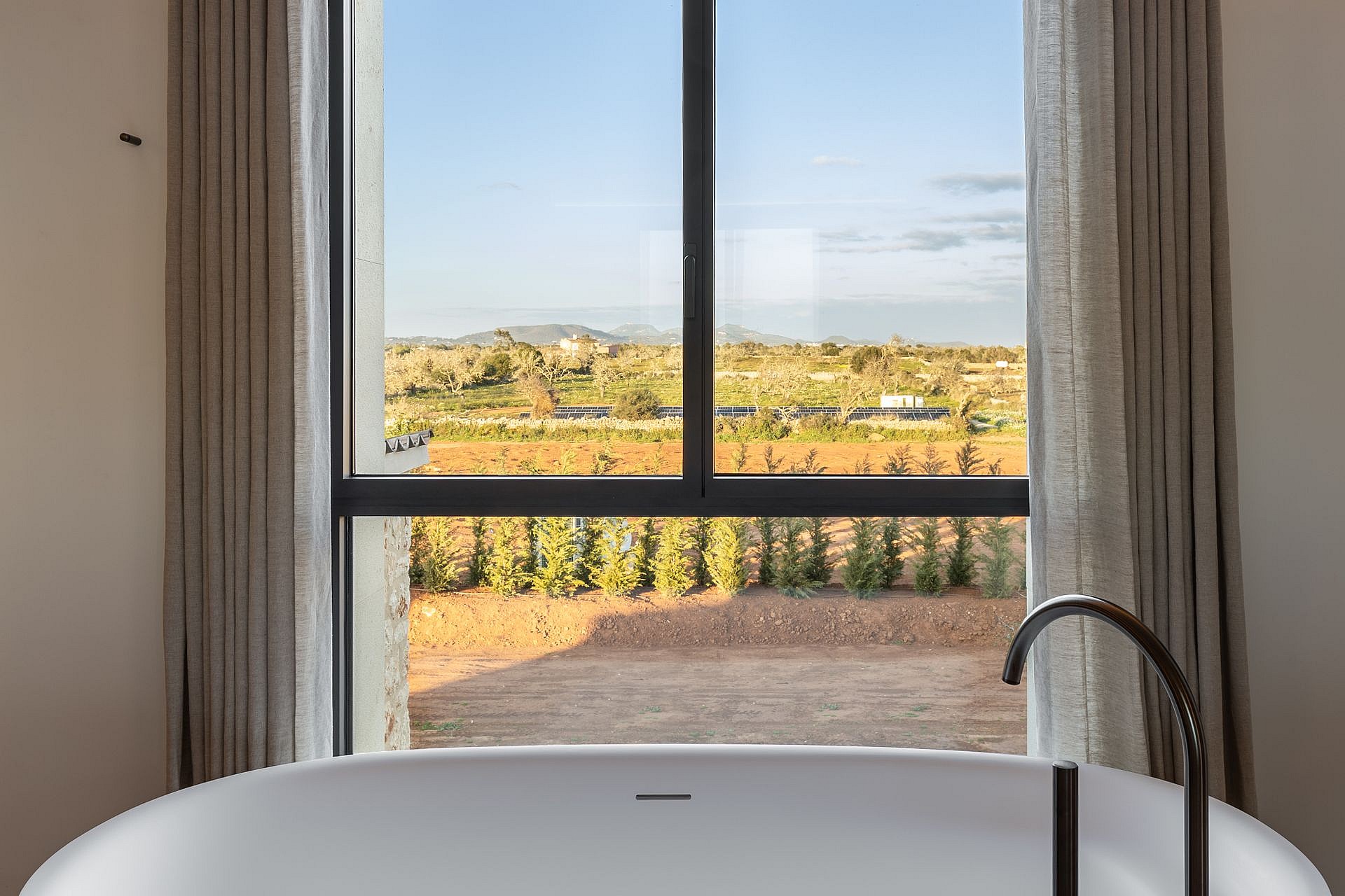 A white freestanding bathtub positioned before a large window looking out onto a rural landscape with distant hills.