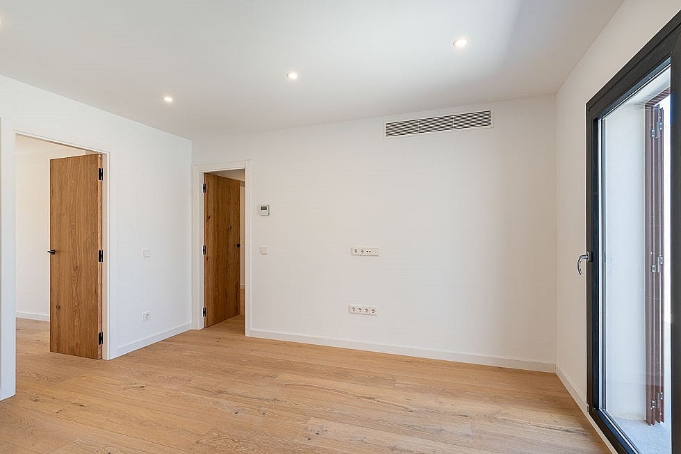 Empty bedroom interior with light wood flooring, white walls, two wooden doors, and a glass terrace door.