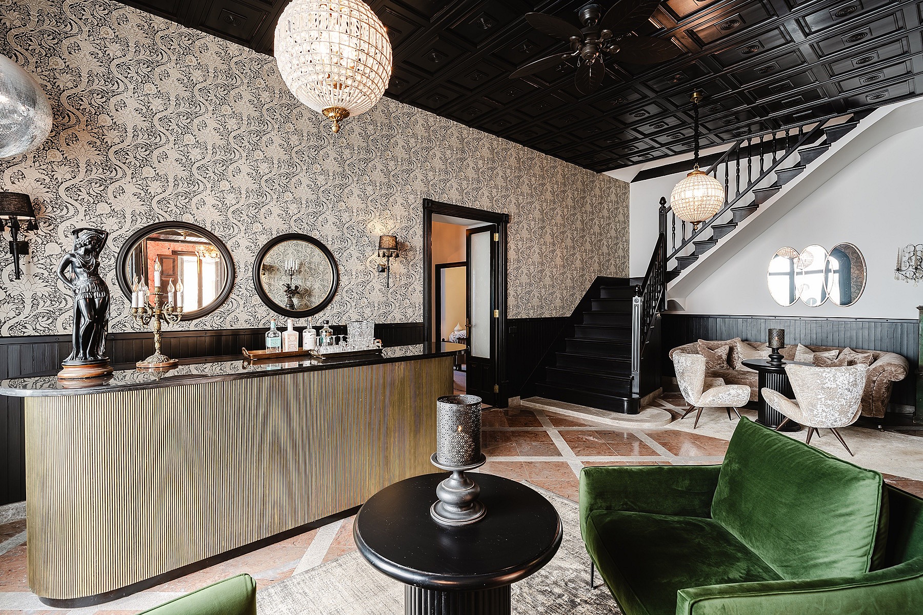 Wide shot of a sophisticated entrance hall featuring a metallic ribbed bar, patterned wallpaper, black ceiling, and green velvet armchairs.