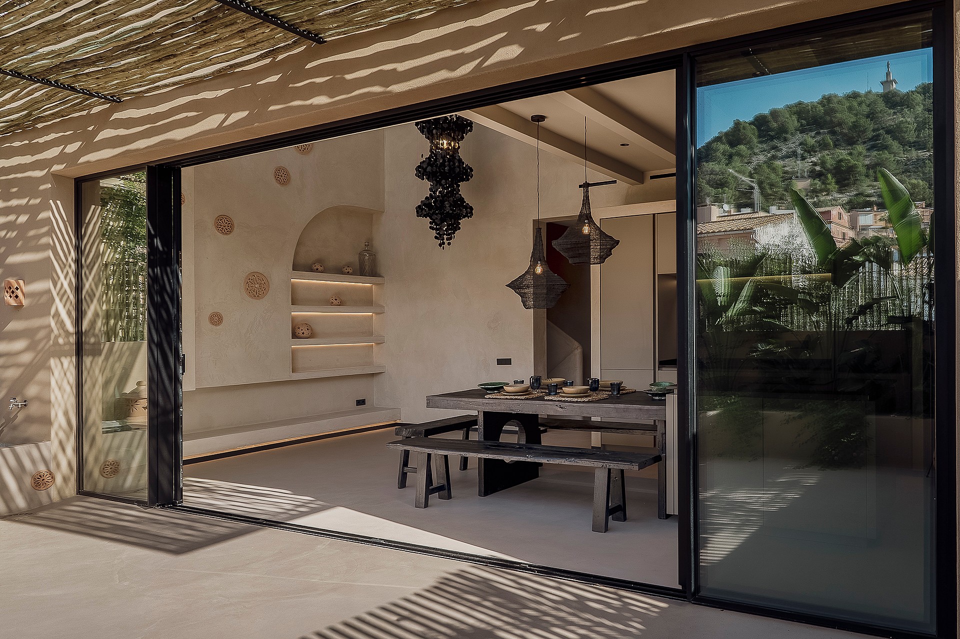 An open-plan dining room with a long wooden table and benches leading onto a terrace shaded by a reed canopy.