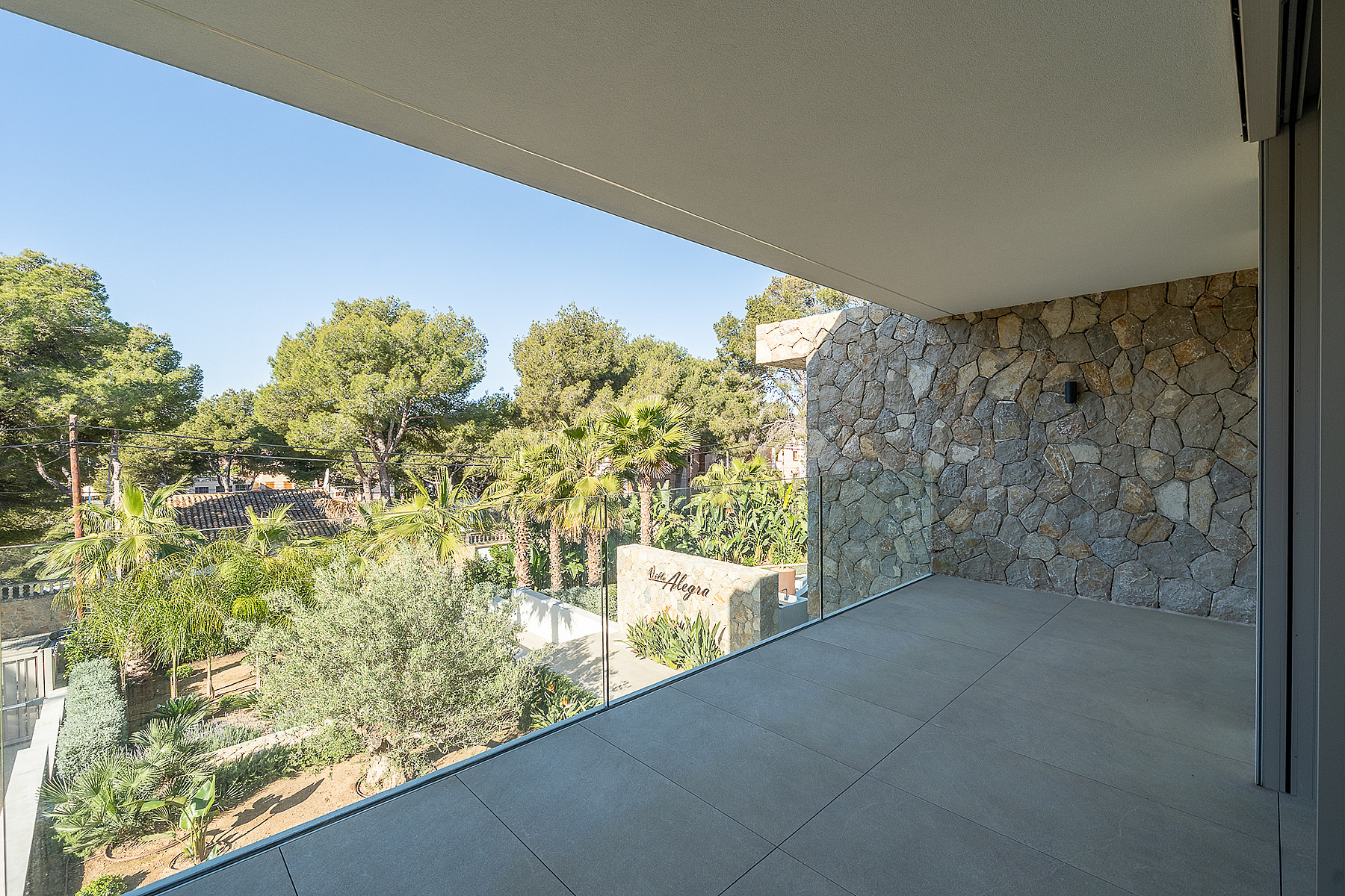 A sheltered terrace with a rough-hewn stone feature wall and glass balustrade, overlooking a Mediterranean garden with pine trees.