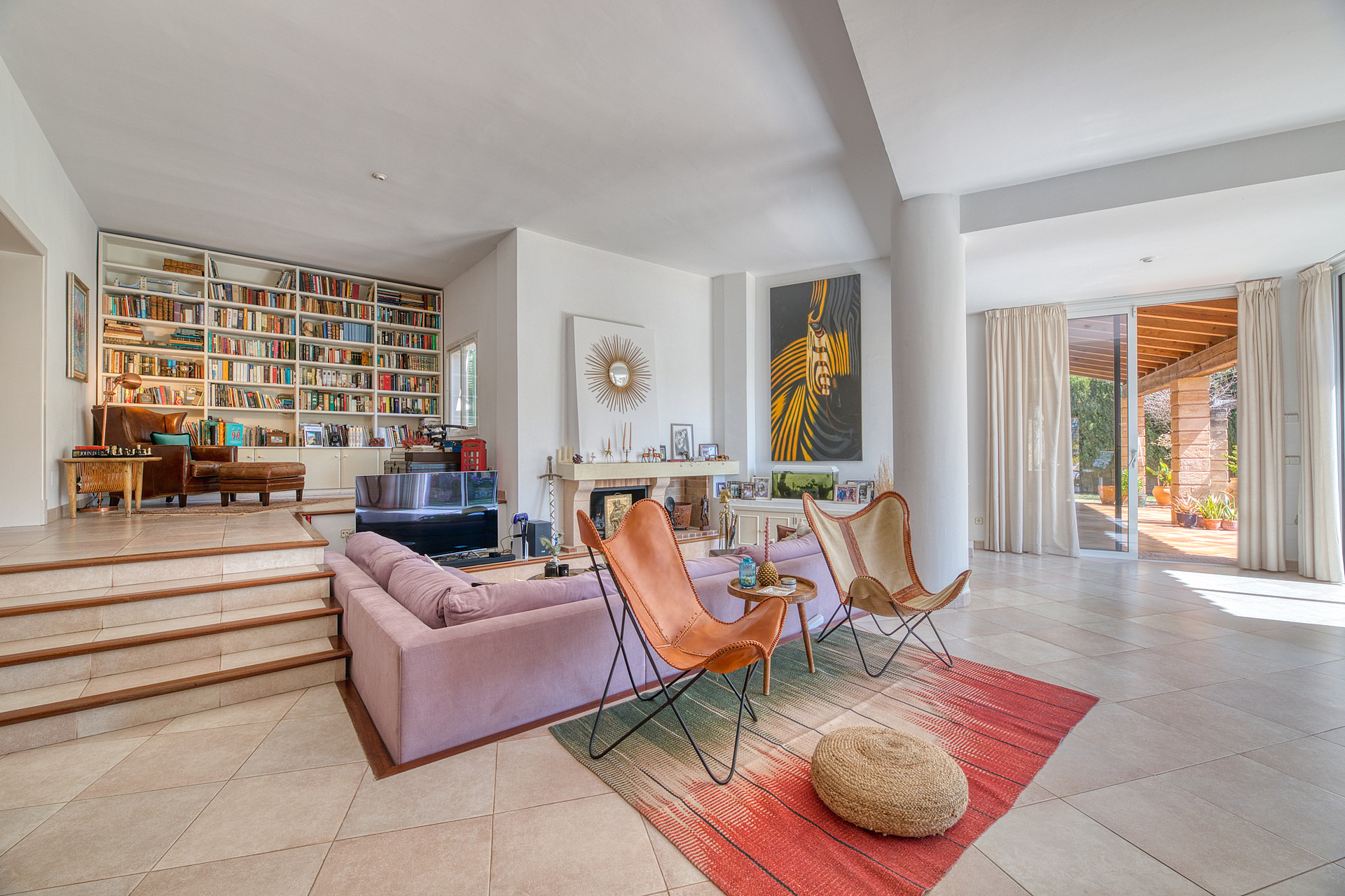 Sunlit living area featuring a split-level layout, large floor-to-ceiling wooden bookshelves, a soft sofa, two leather butterfly chairs, and large glass doors leading to a covered terrace.