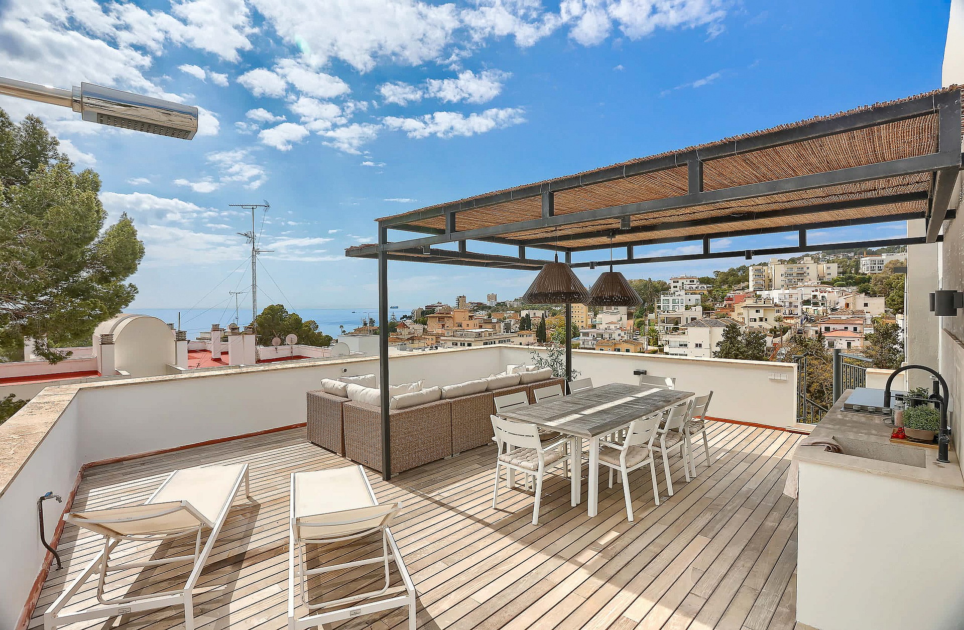 A spacious outdoor rooftop terrace featuring a wooden deck, a covered dining area with a woven pergola, lounge seating, and distant sea views over a coastal neighborhood.