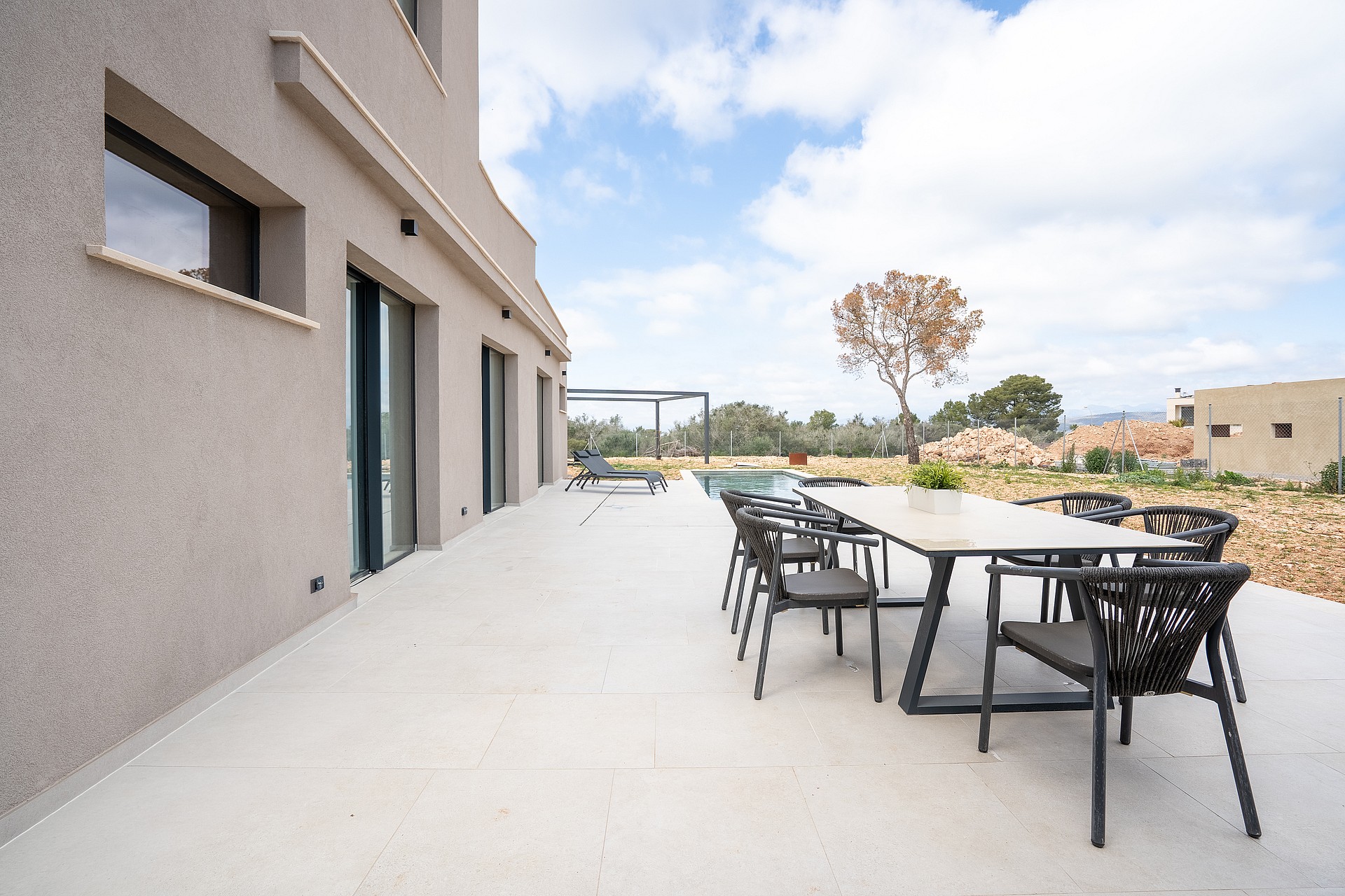 Outdoor dining set on a pale concrete terrace beside a villa wall, overlooking distant mountains under a cloudy sky.