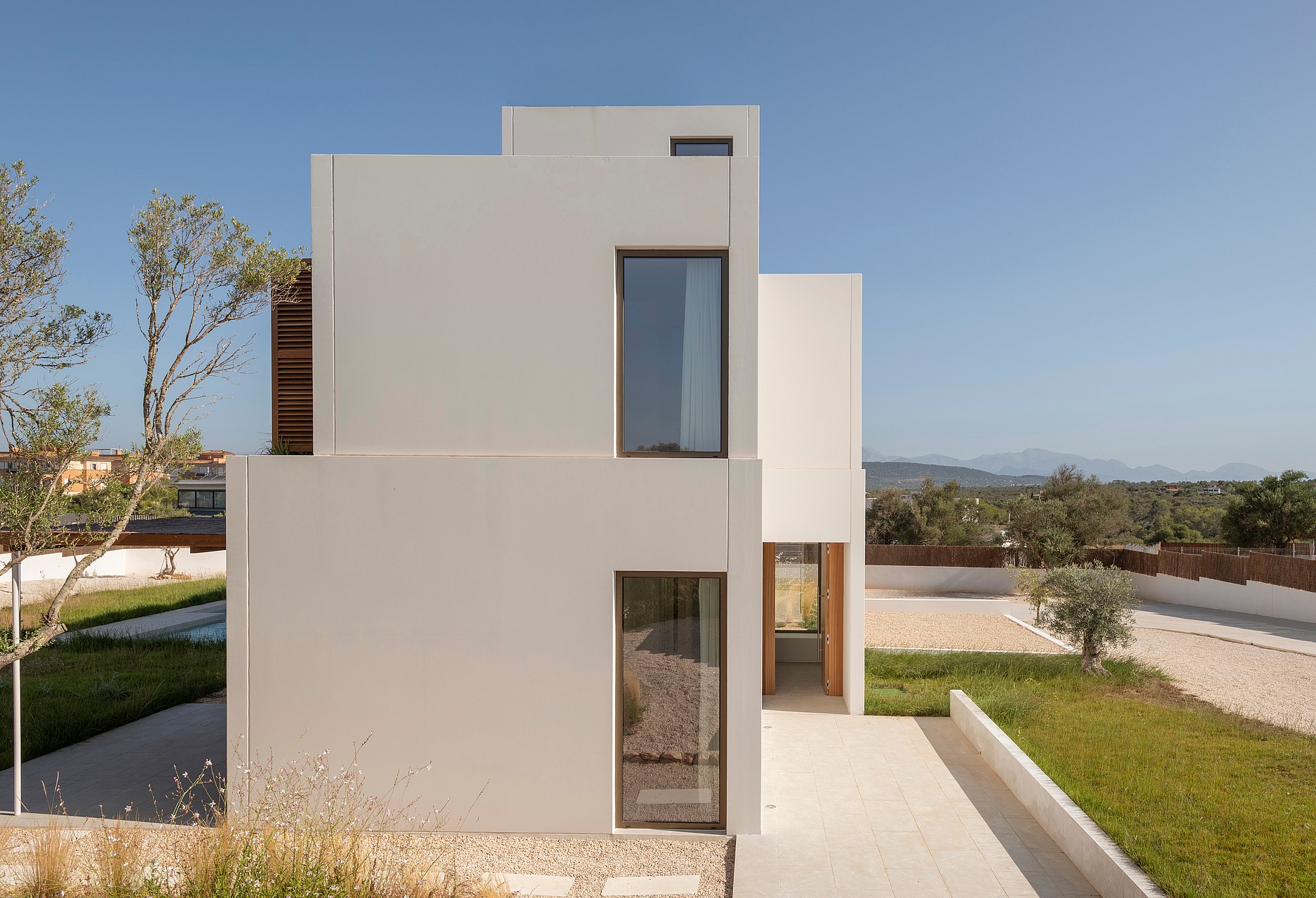 Modern two-story villa featuring white geometric volumes, vertical windows, and wooden shutters under a clear blue sky.