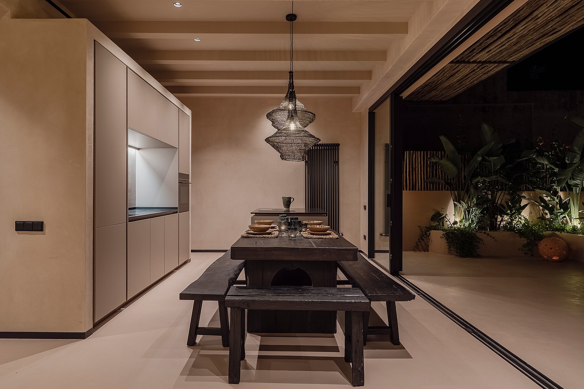 Interior dining space featuring a dark wood table and benches, illuminated by a modern pendant light, opening to an outdoor terrace at night.