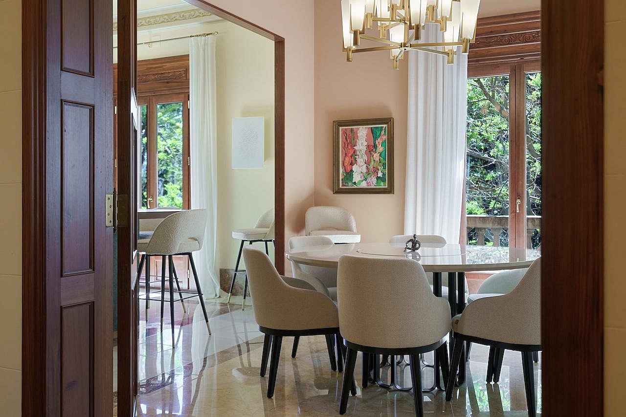 Dining room with a round marble table, upholstered chairs, a crystal chandelier, and large windows overlooking trees.