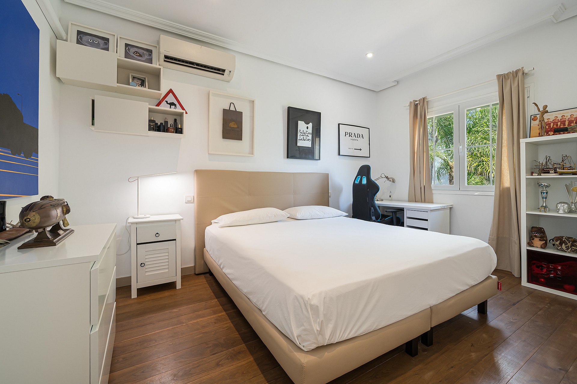 A bright bedroom with a queen-sized bed, white linens, a wooden floor, and a small workspace. A window looks out onto green foliage.