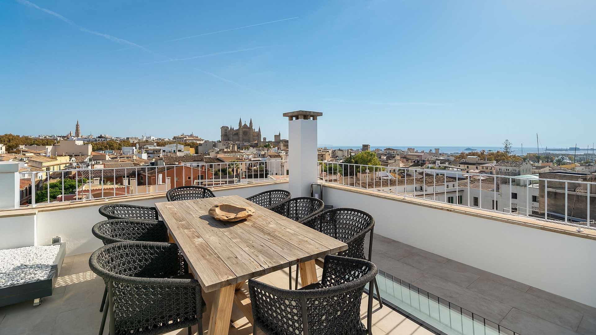 Rooftop terrace with wooden dining table and black chairs overlooking Palma Cathedral and the Mediterranean sea.