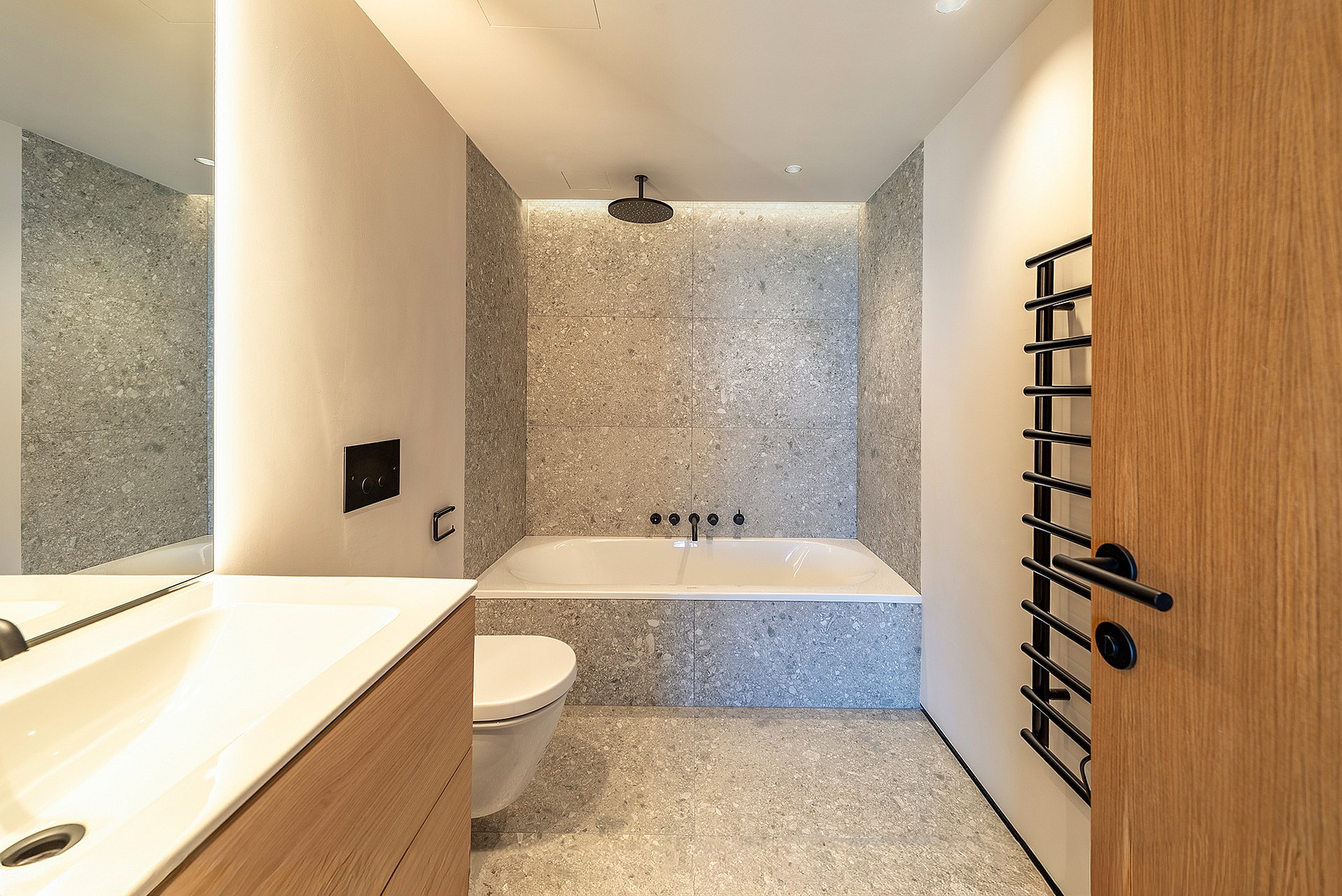 A modern bathroom featuring a white bathtub encased in grey mosaic tiles, a wooden vanity, and a black heated towel rail.