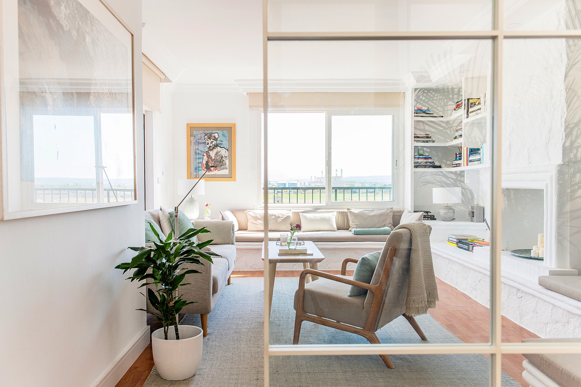 A view through a glass partition into a bright living room with a neutral sofa, patterned area rug, and an armchair.