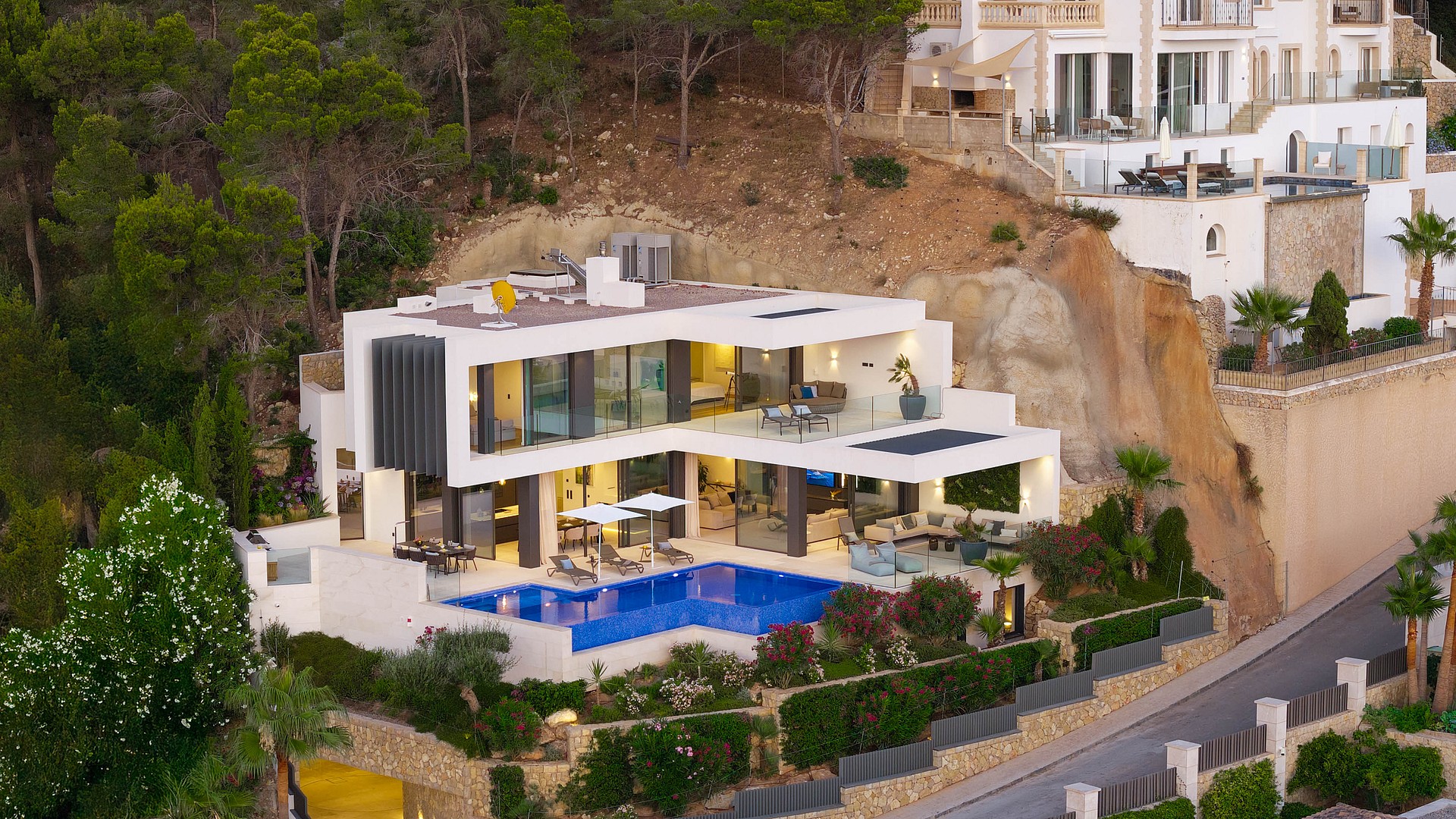 Aerial view of a white modern villa carved into a cliffside, featuring a swimming pool, multiple terraces, and glass facade.
