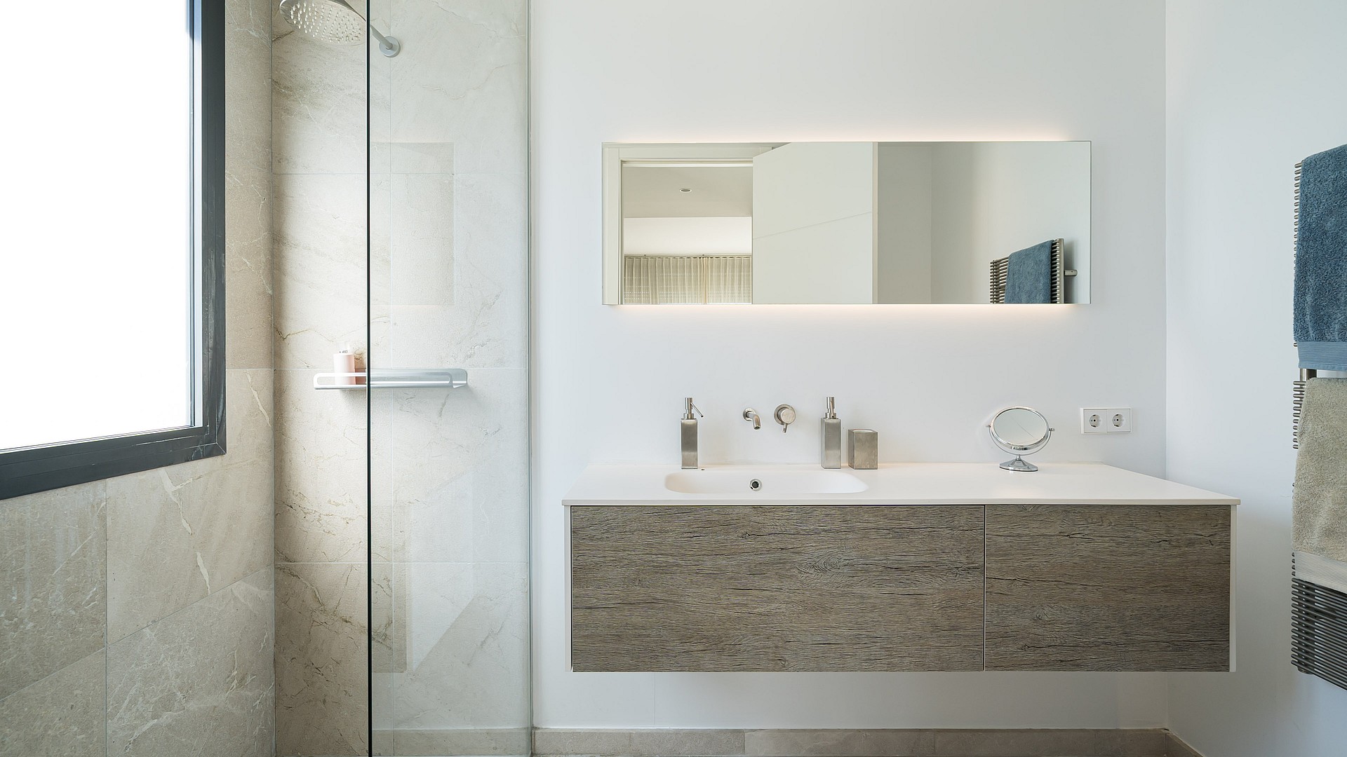 A modern bathroom featuring a glass-enclosed shower with light-toned stone tiling, a floating vanity with a wooden cabinet, and a wall-mounted mirror.