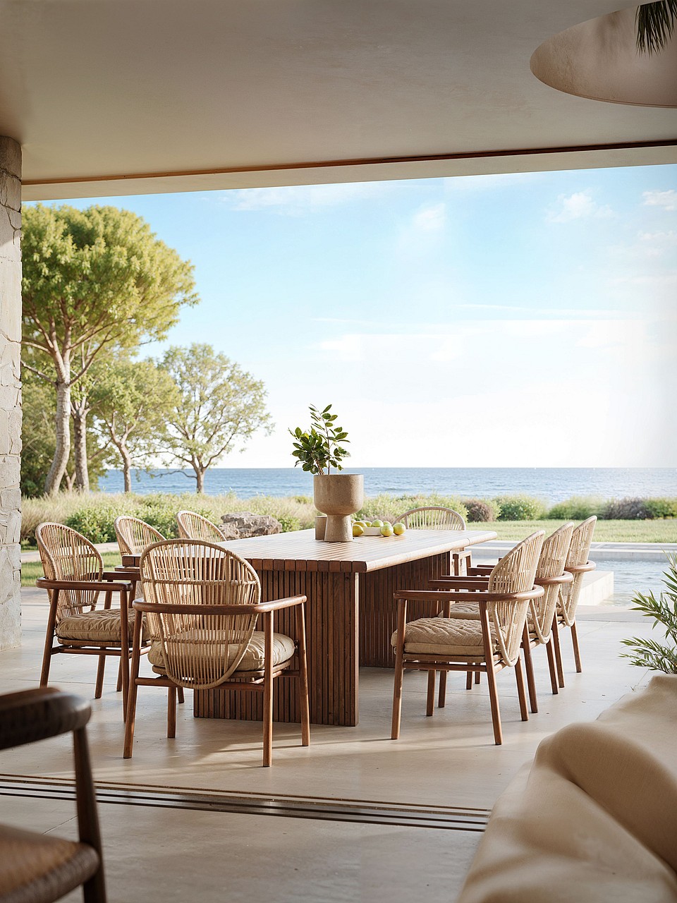 Outdoor dining set with a long wooden table and woven chairs on a covered terrace, overlooking the sea and garden.