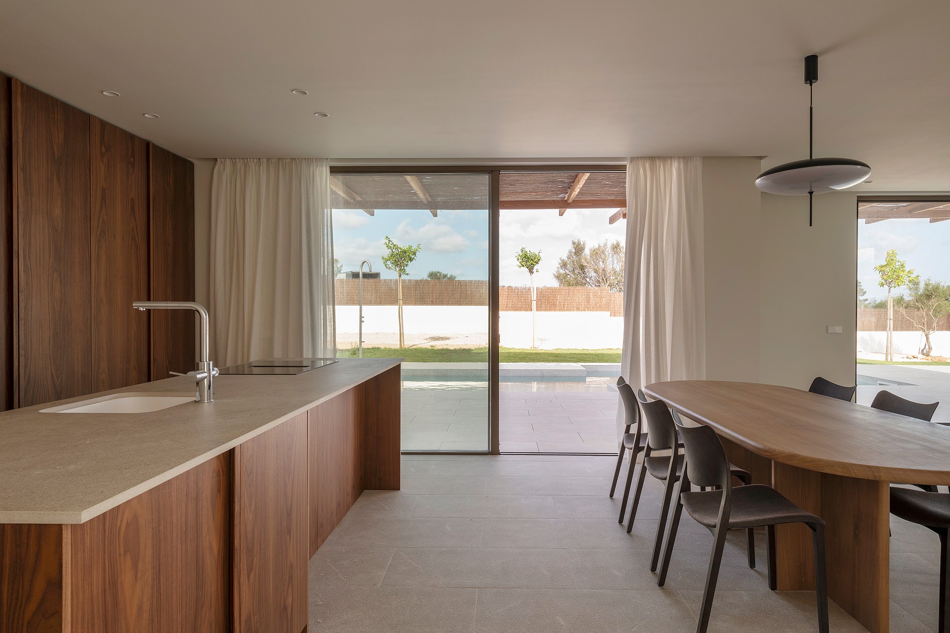 An oval oak dining table and dark chairs in a sunlit room with stone floors and floor-to-ceiling glass sliding doors.