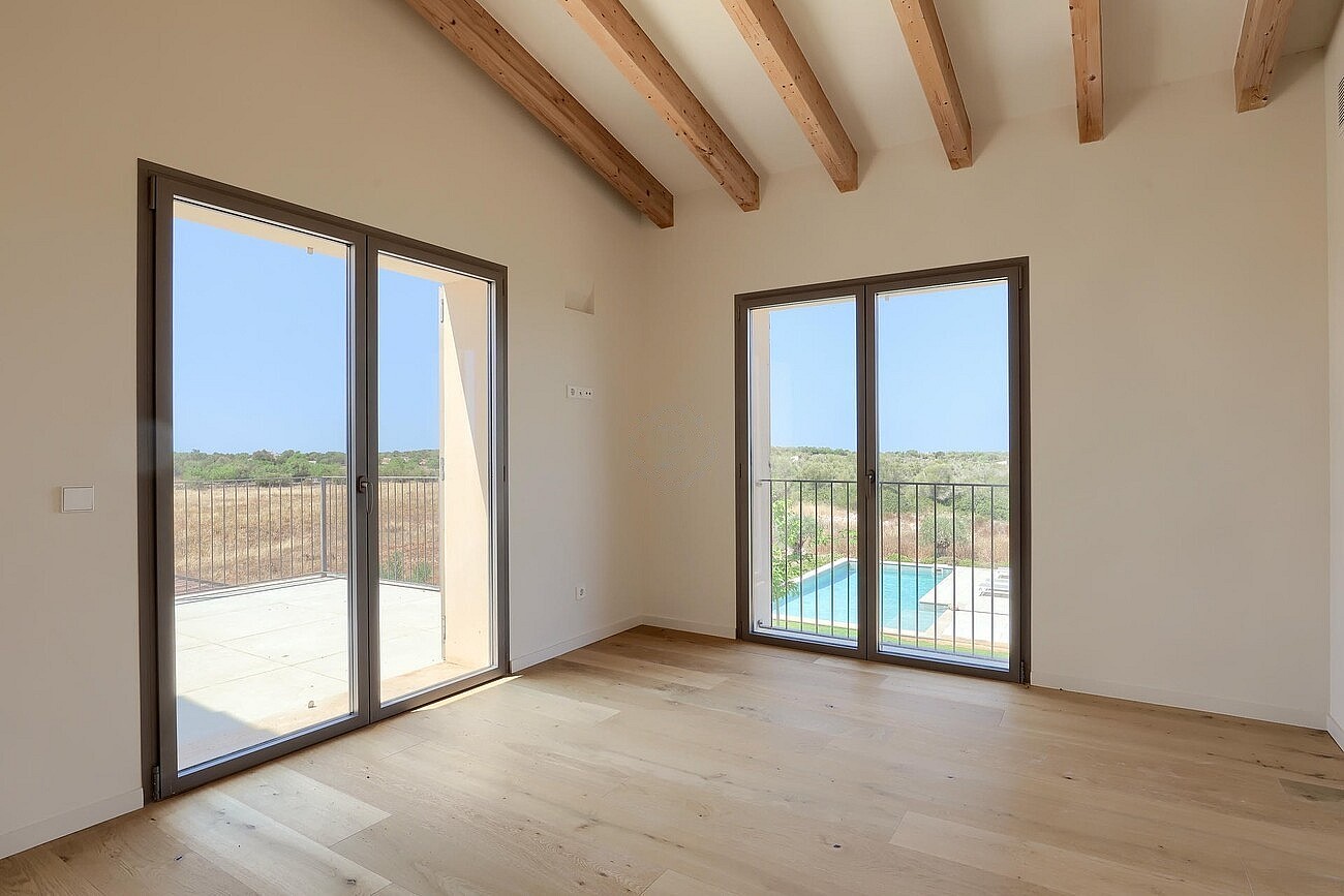 An unfurnished bedroom in a Finca, featuring light oak wood floors, white walls, exposed wooden ceiling beams, and sliding glass doors opening to a view of the pool and surrounding landscape.
